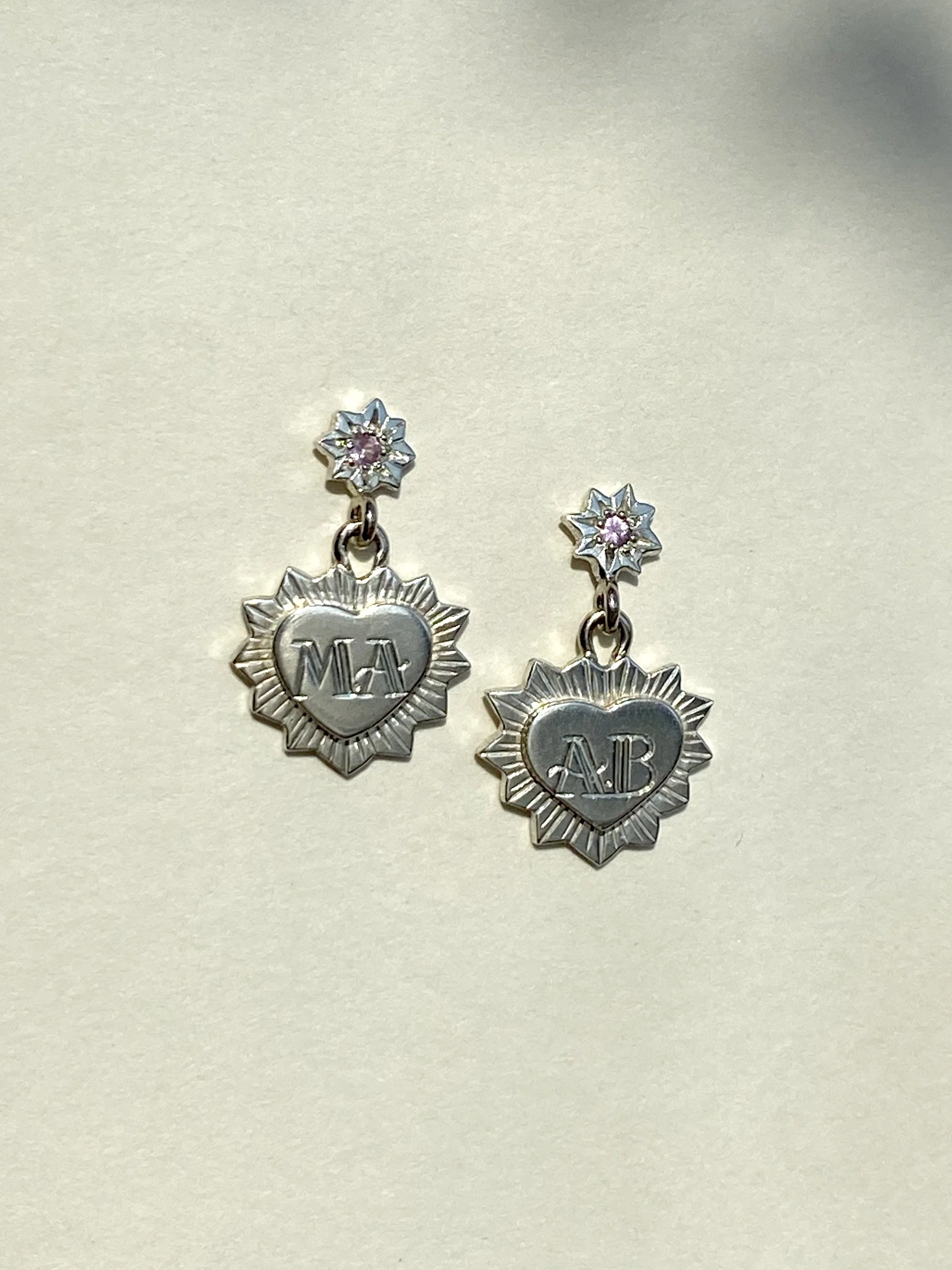 White gold and pink sapphire hand engraved earrings with a star-shaped top and a heart-shaped pendant engraved with 'MA' on the left and 'AB' on the right.