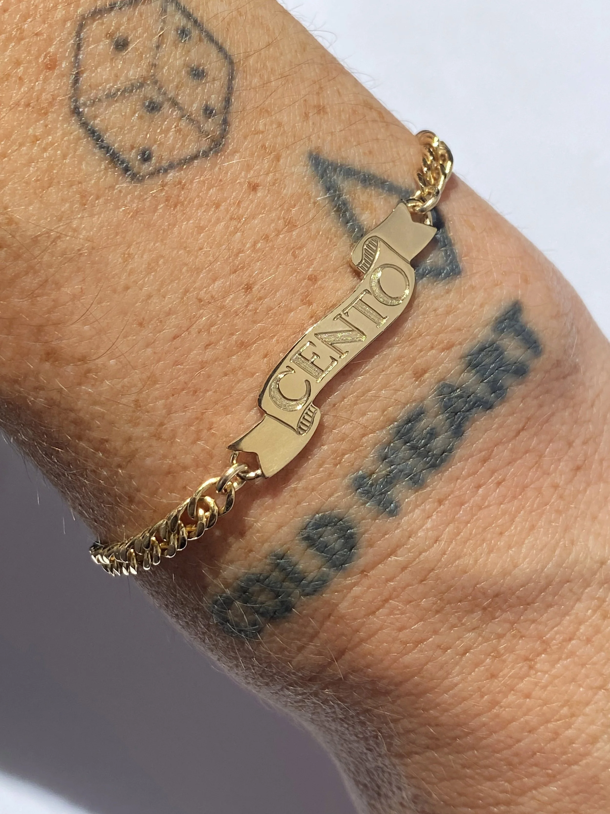 A close-up of a wrist with tattoos and a gold bracelet with a charm that spells 'CELINE'.