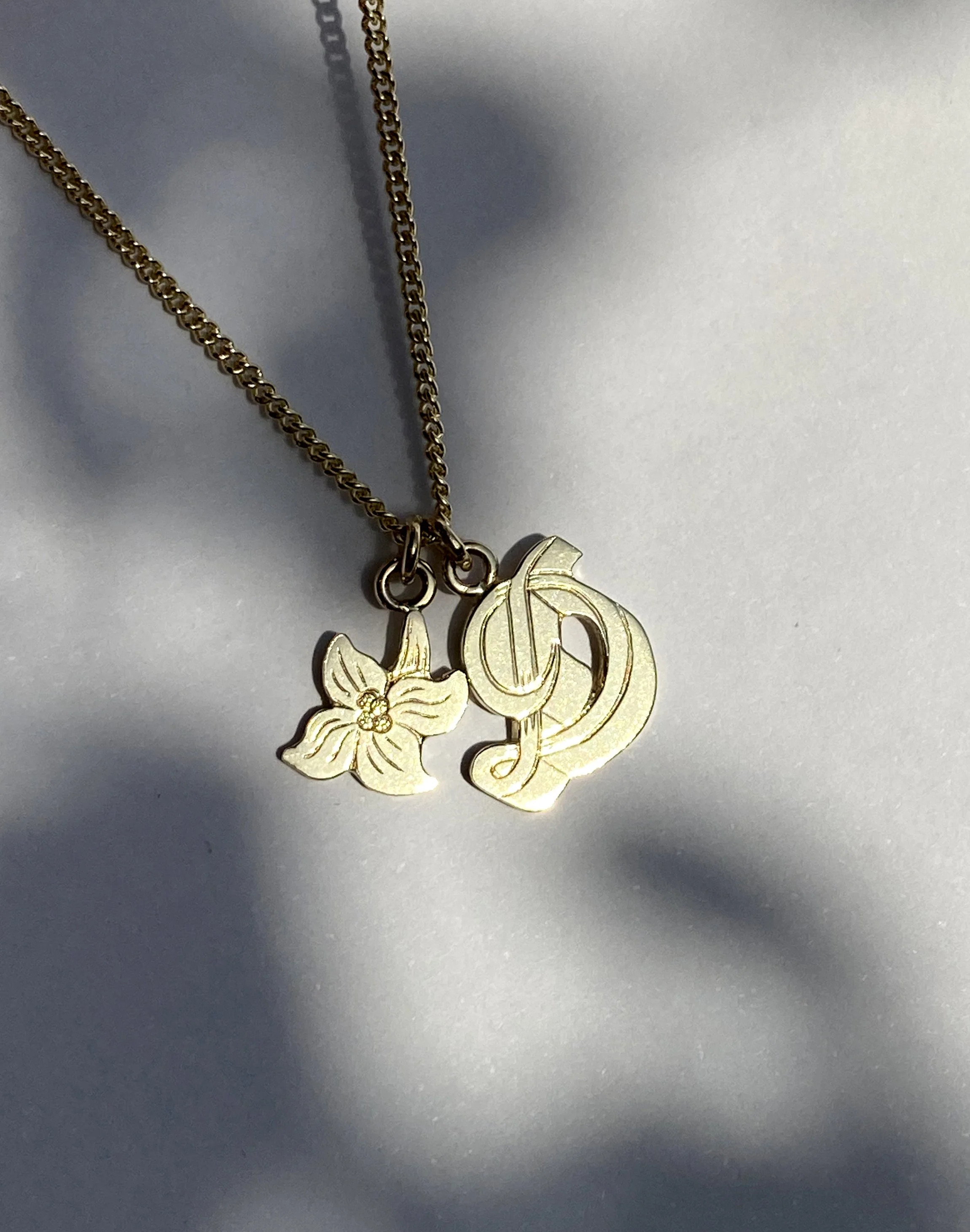 Gold necklace with two charms: a flower and a stylized letter 'D'.