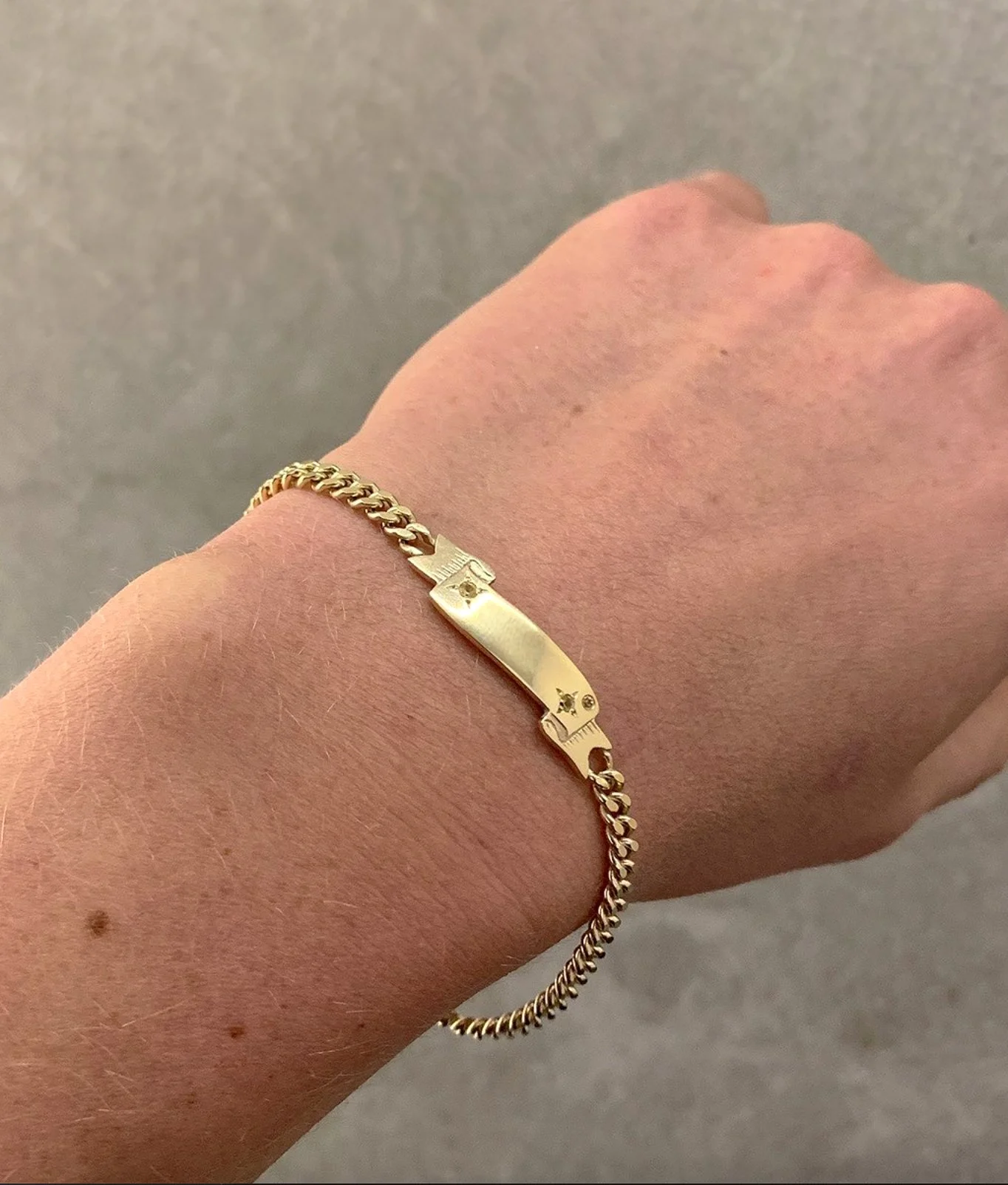 A close-up of a person's wrist displaying a gold bracelet with a rectangular plate in the center, engraved with small decorative elements, against a plain gray background.