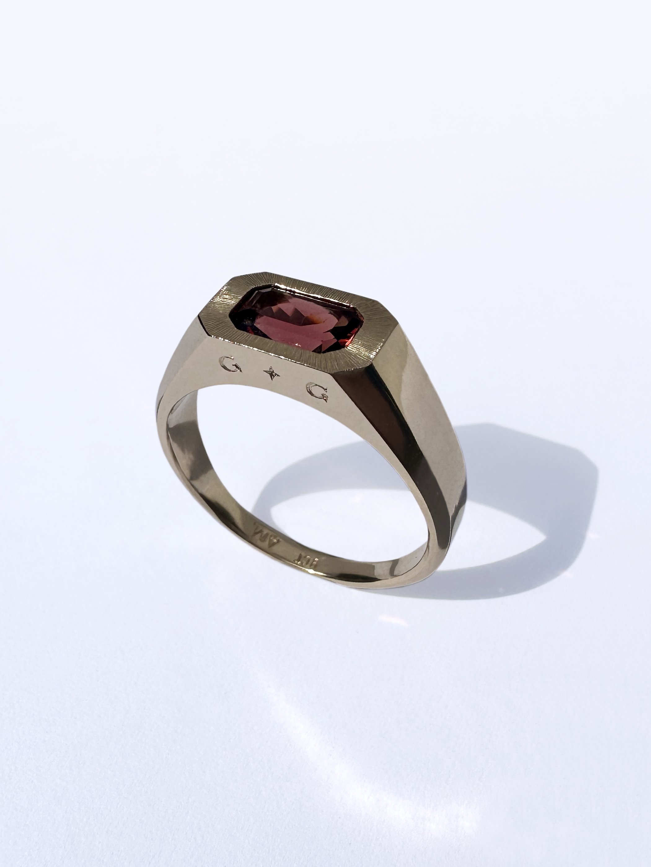 A gold ring with a rectangular red gemstone set in a flat bezel, featuring engraved letters around the band.