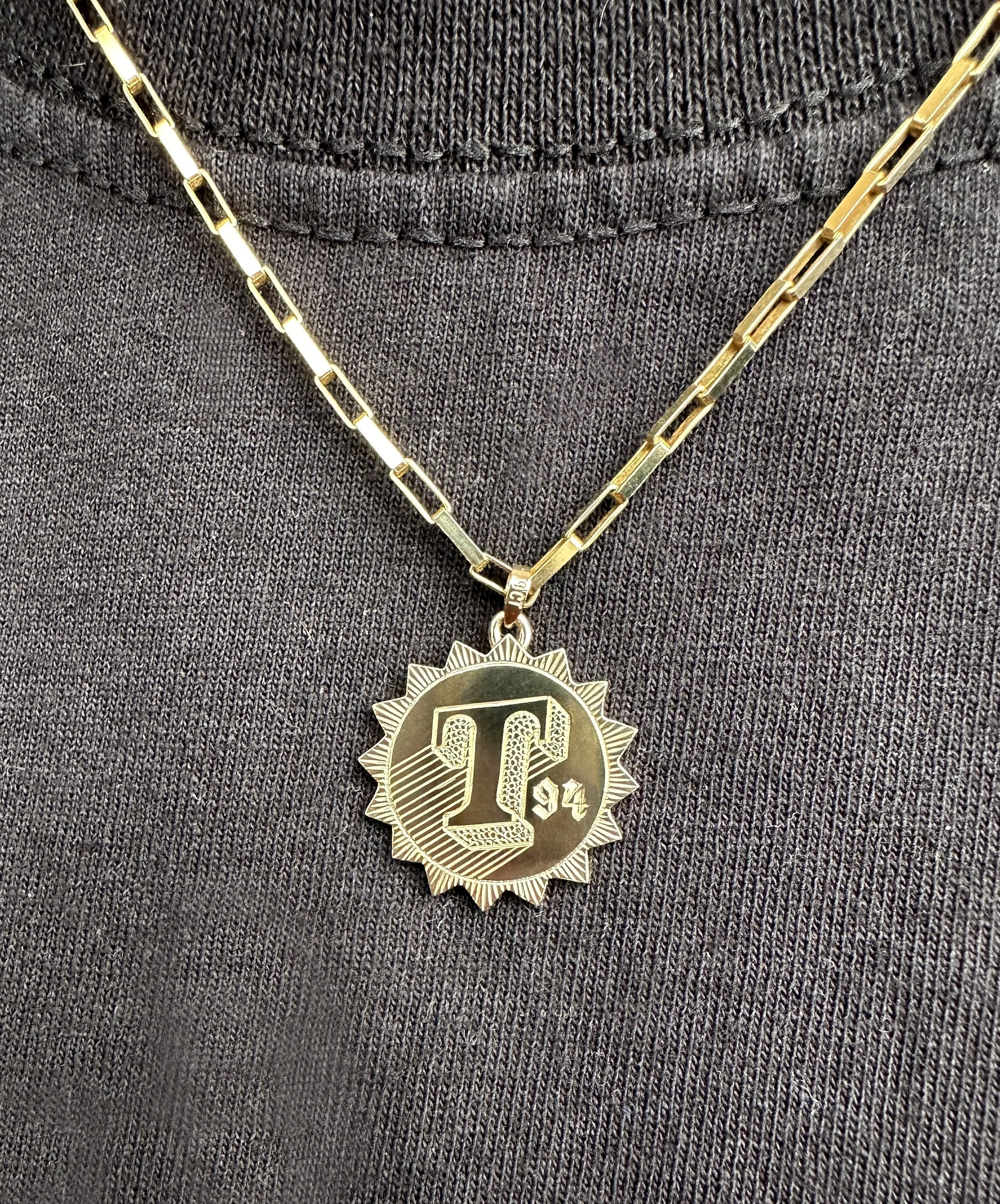 Gold chain necklace with a pendant featuring the letter 'L' and the year '94' on a dark fabric background.