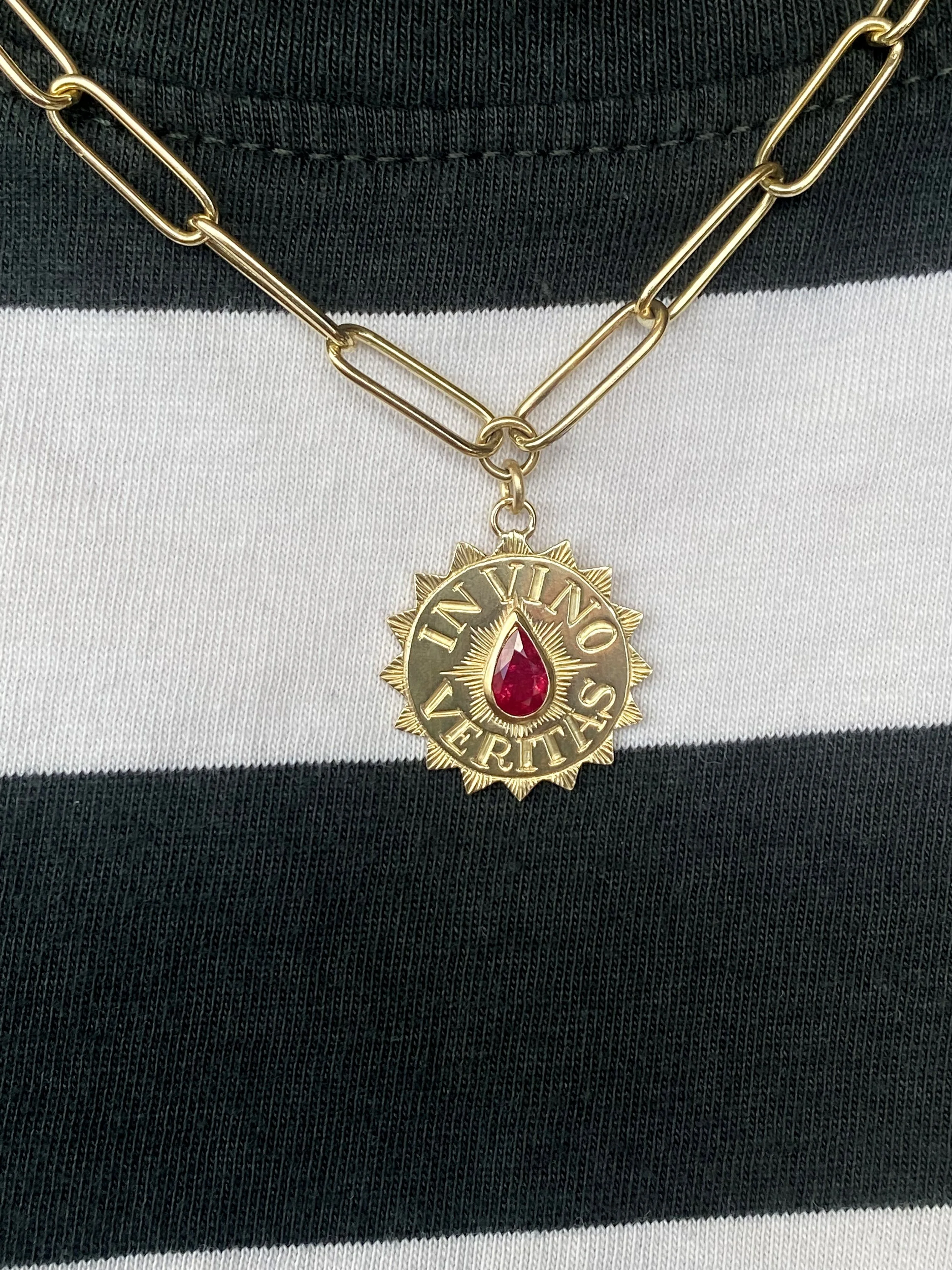 Gold-tone necklace with a pendant featuring a red teardrop gem and the words 'IN VINO VERITAS' on a black and white striped fabric background.