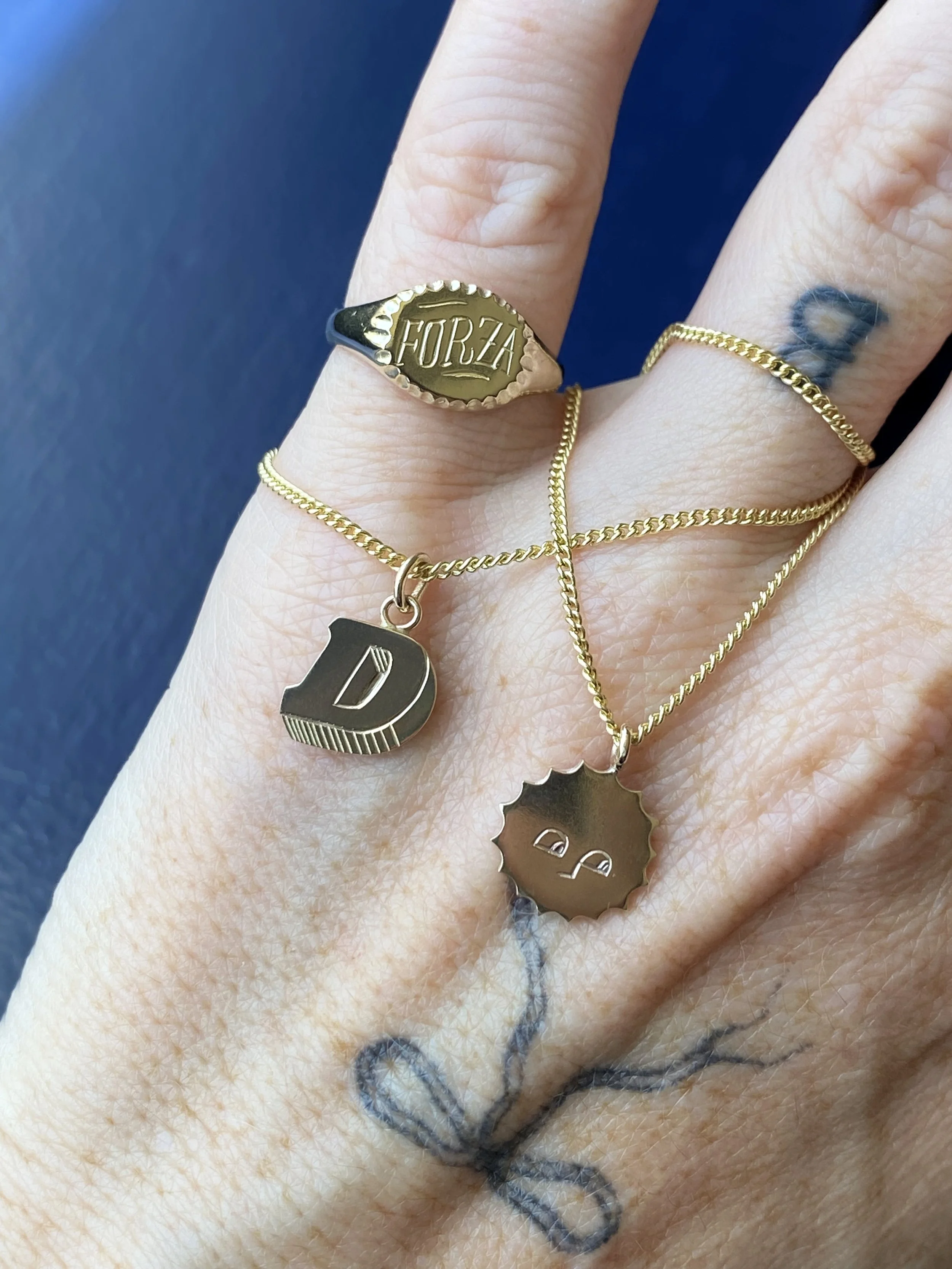 Multiple gold rings on fingers and two necklaces with charms, one with a letter D and the other with a face, resting on a person's hand with a black tattoo of a plant or tree.