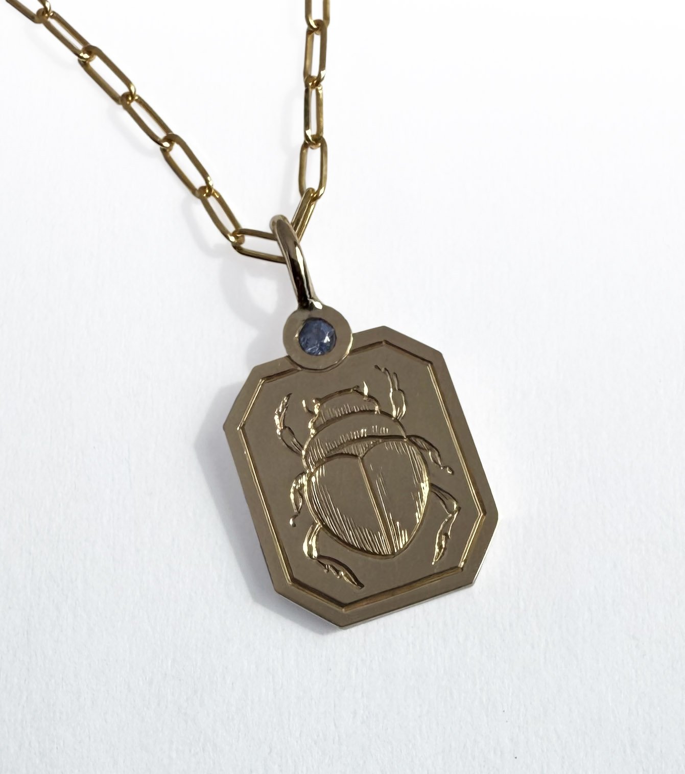 A gold necklace with a pendant shaped like a beetle, engraved on a rectangular background, with a small gemstone on the bail, set against a white background.