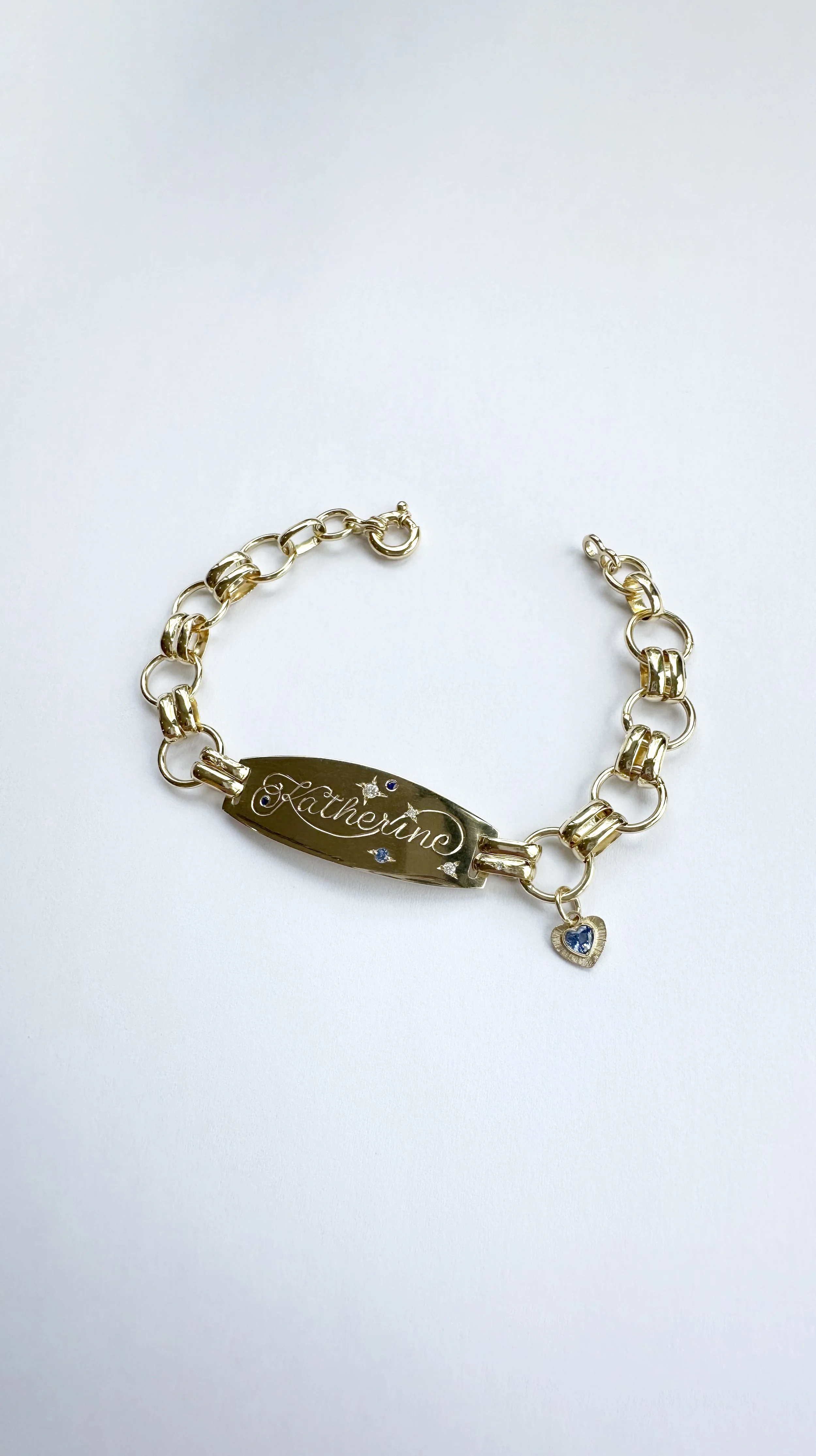 Gold bracelet with a central engraved plate that says 'Katherine' and has small blue gemstone accents, along with a small heart-shaped charm with a blue gemstone.