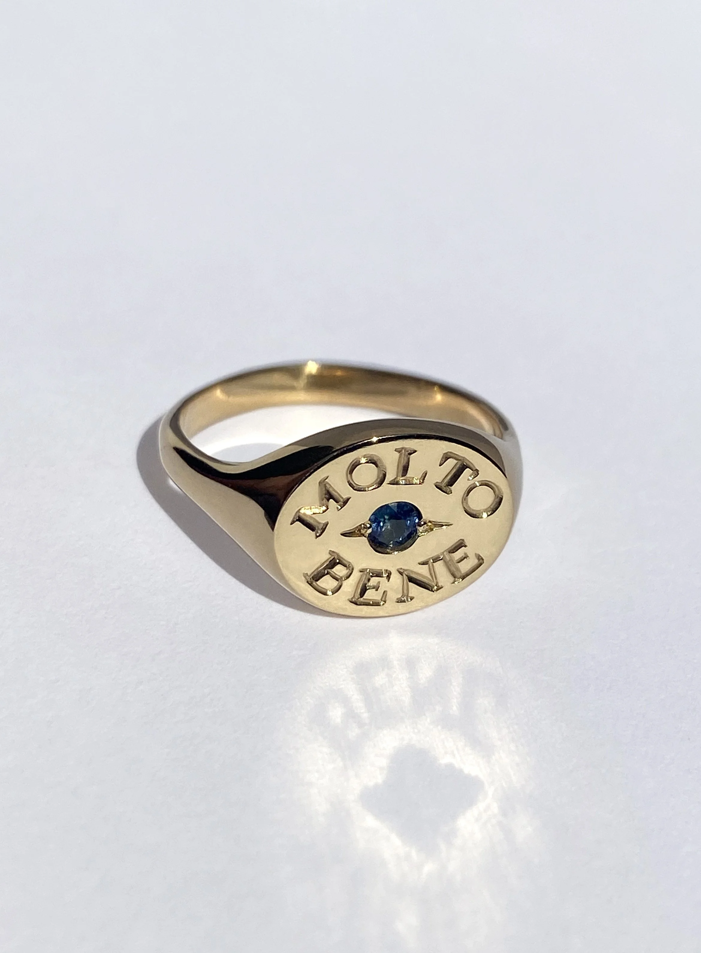 18k gold ring with a circular face engraved with 'MOLTO BENE' and a small blue Australian sapphire in the centre.