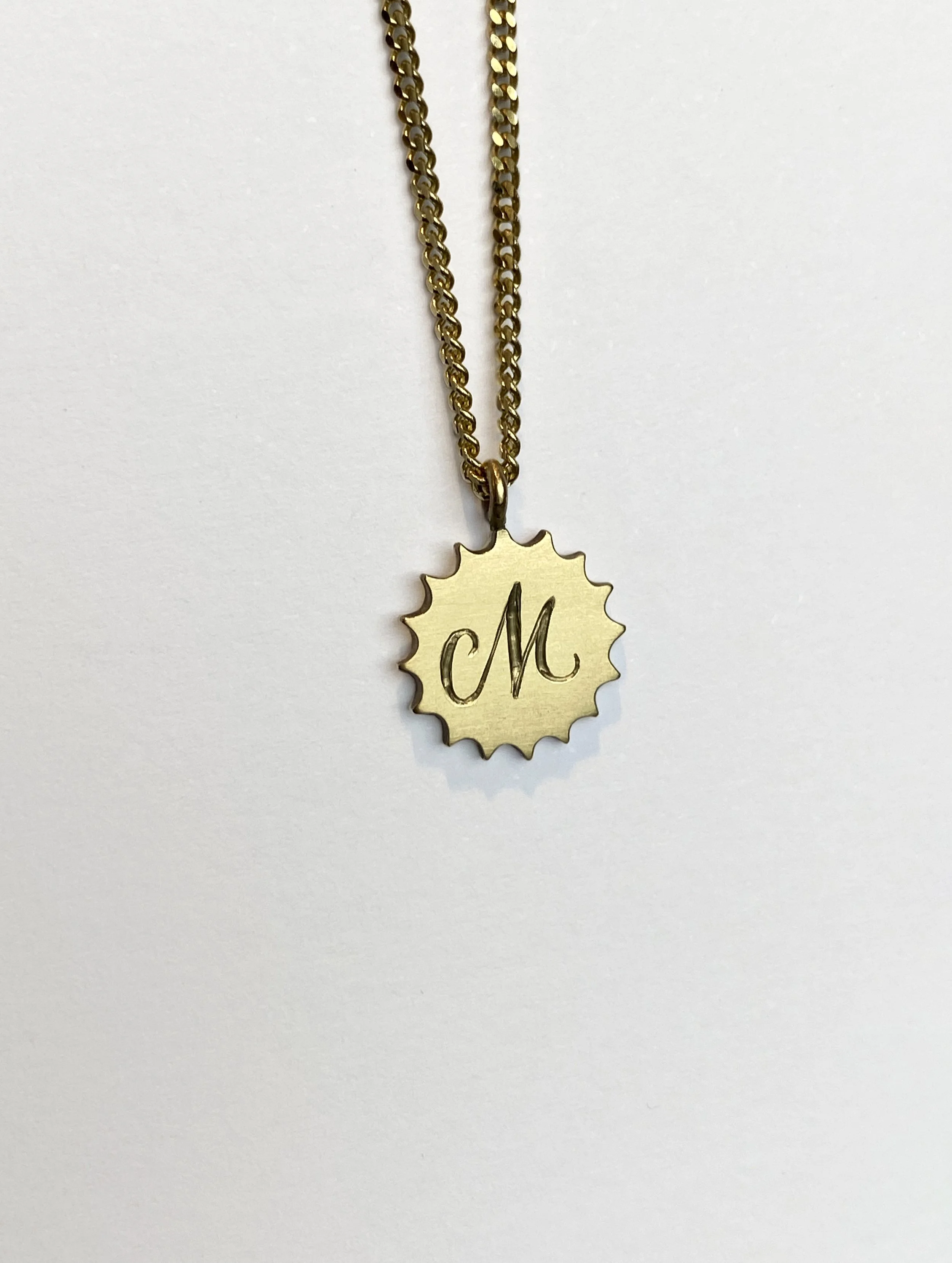 Gold necklace with round pendant engraved with a stylized letter 'M' against a plain white background.