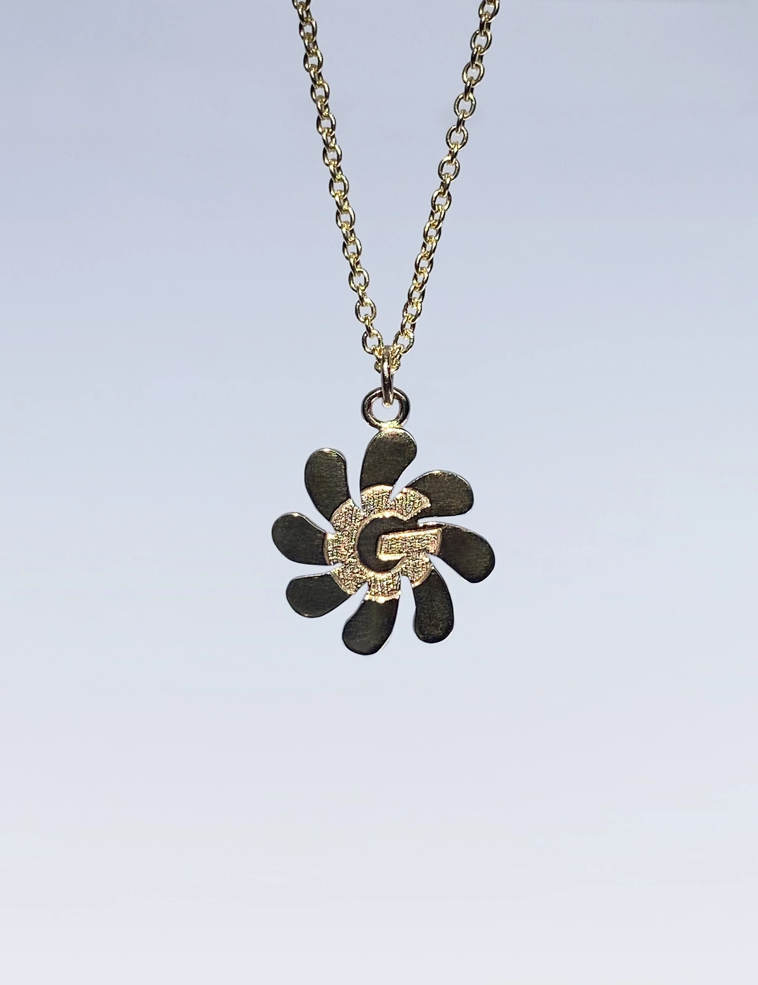 Gold necklace with a floral-shaped pendant featuring a letter 'G' at the center, hanging against a plain background.