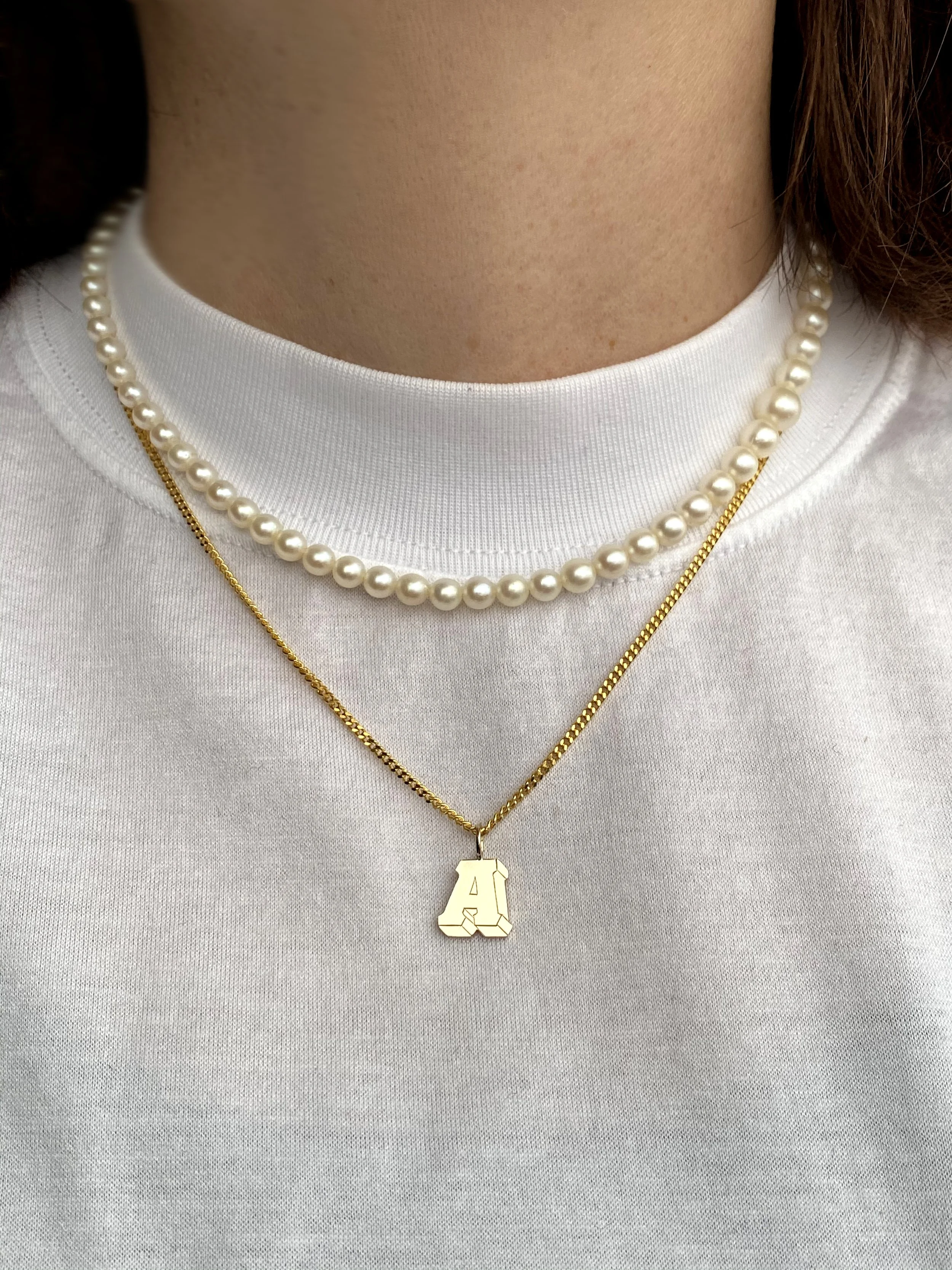 Close-up of a person's neck wearing a pearl necklace, a gold chain necklace with a letter 'A' pendant, and a white crew-neck shirt.