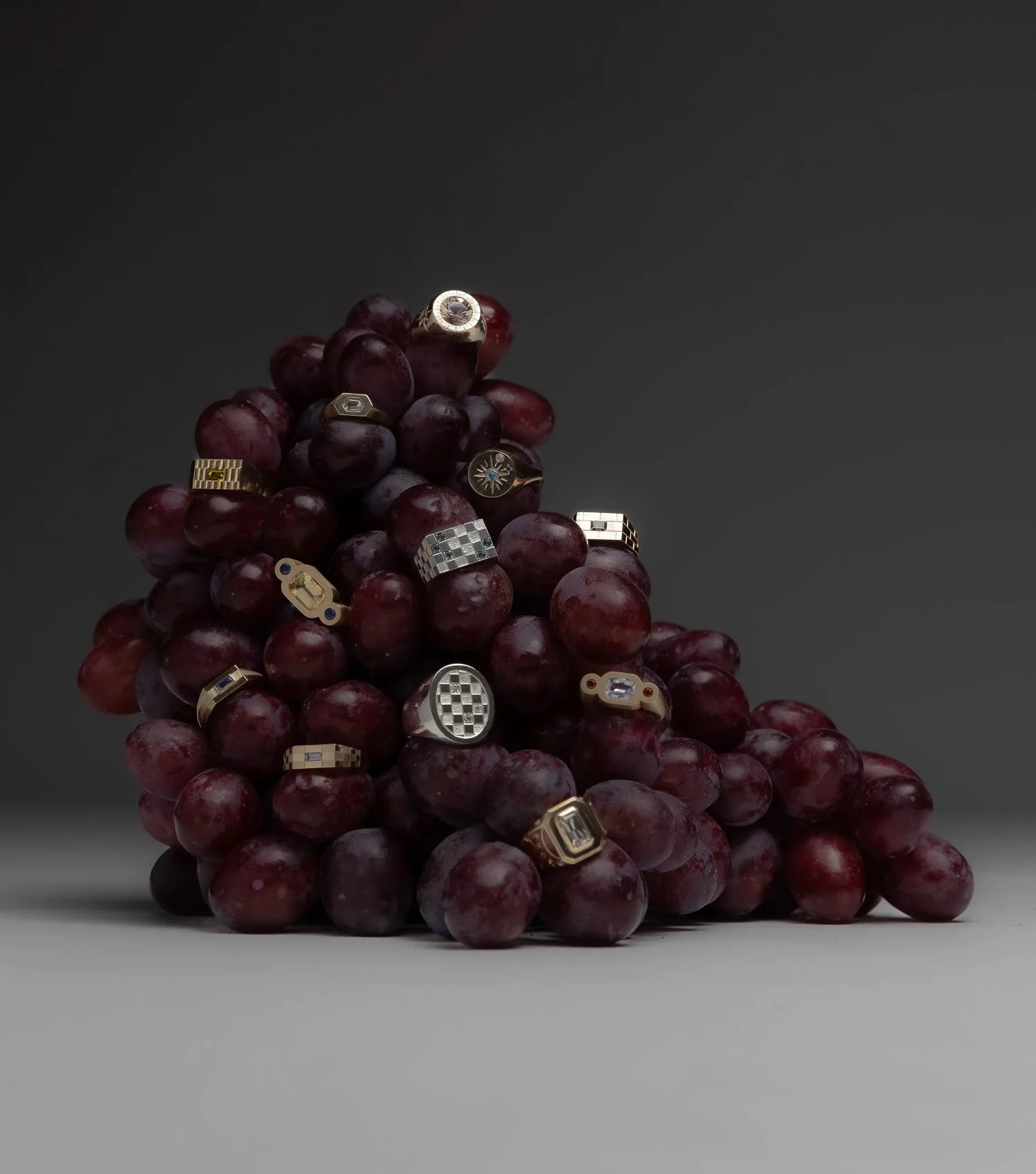 A bunch of red grapes with various gold and silver rings and jewelry pieces scattered throughout. Photographed by Holly Graham