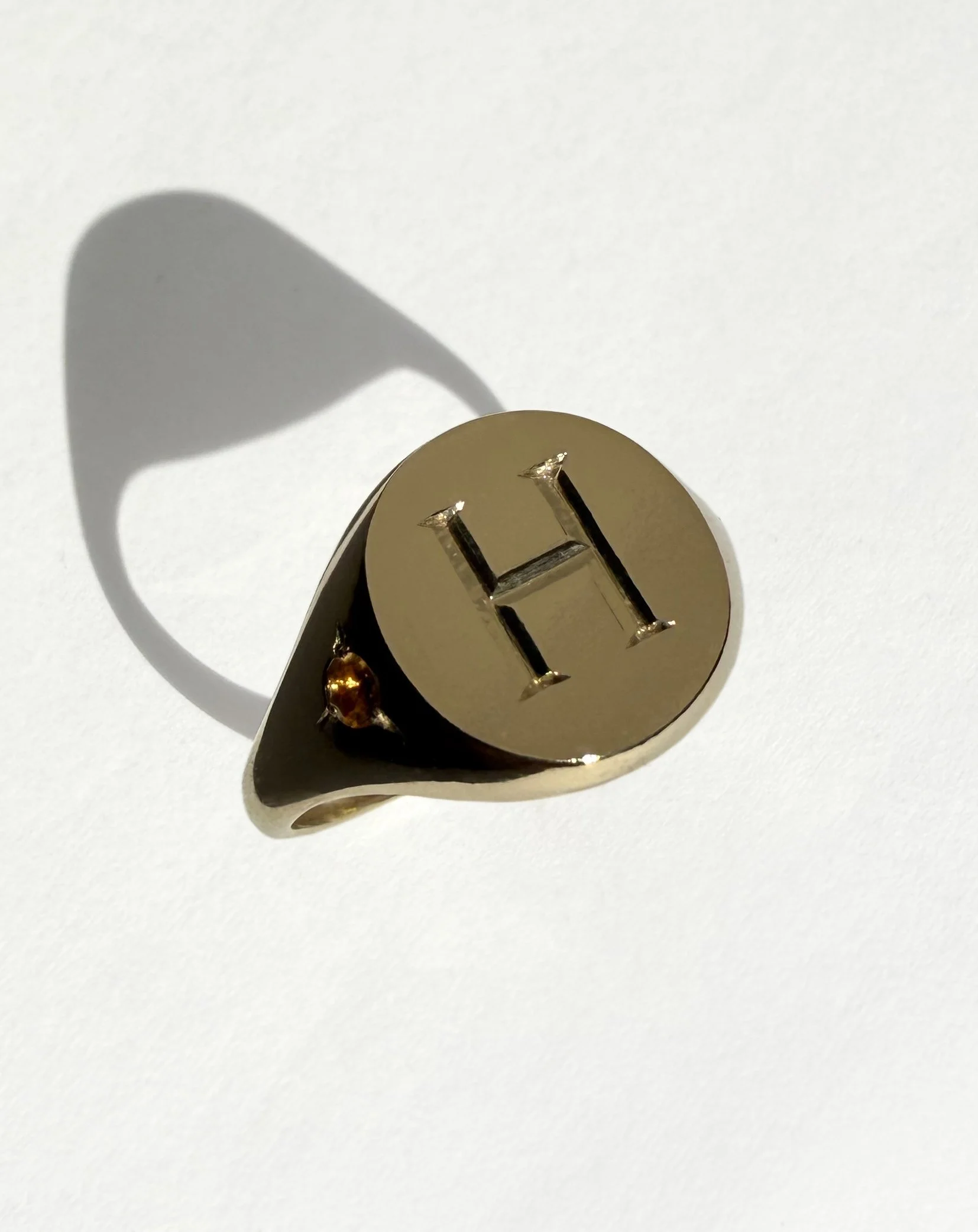 Gold ring with engraved letter 'H', featuring a yellow stone on the band.