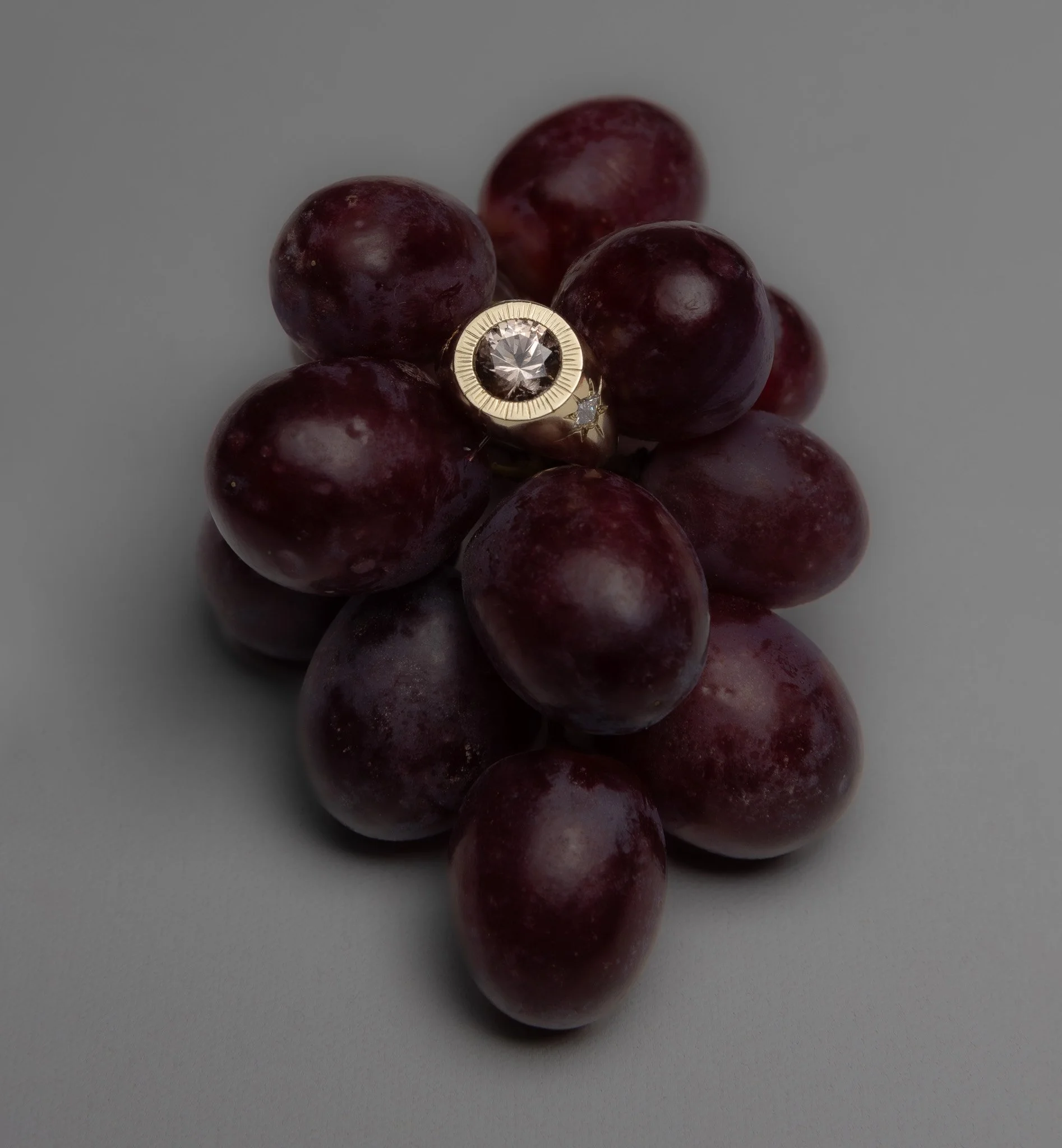 A bunch of red grapes with a 18K gold ring featuring a large 2 carat Parti Sapphire and smaller accent diamonds placed on top. Photographed by Holly Graham