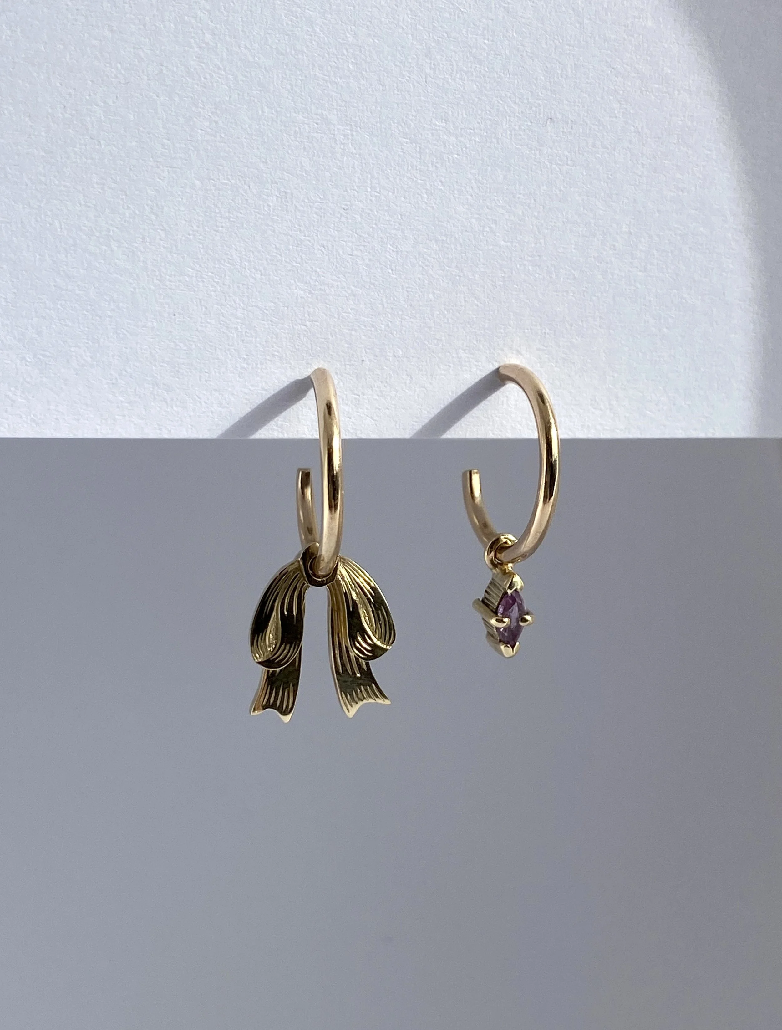 Pair of gold earrings, one engraved with a bow charm and the other with a purple gemstone.