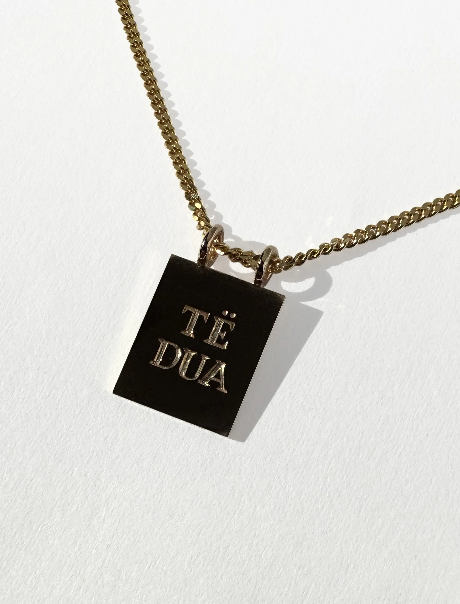 Gold chain necklace with a black pendant engraved with the phrase 'TÉ DUA' in silver letters.
