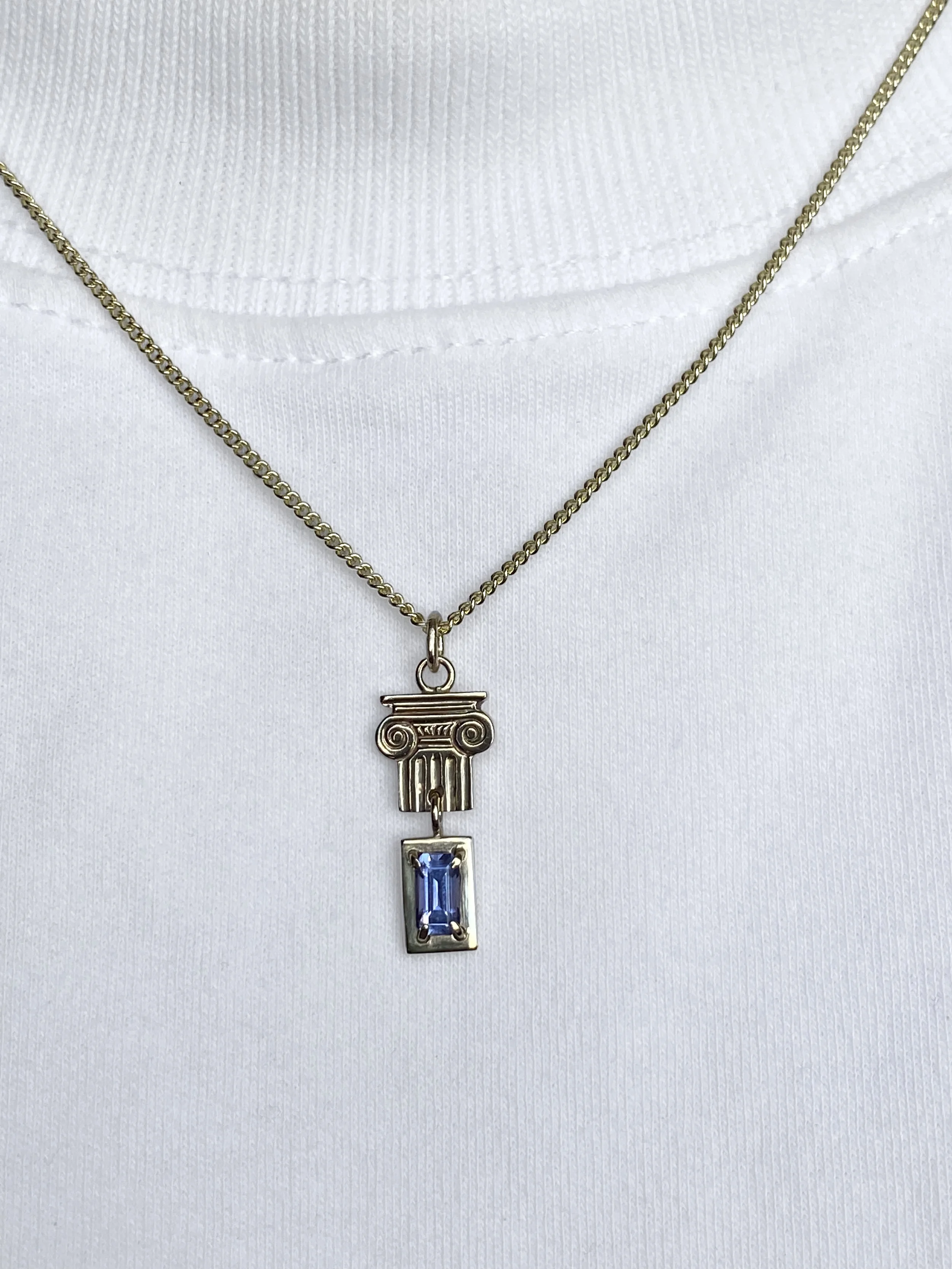 Gold necklace with a pendant featuring a blue rectangular gemstone, hanging from a white fabric background.