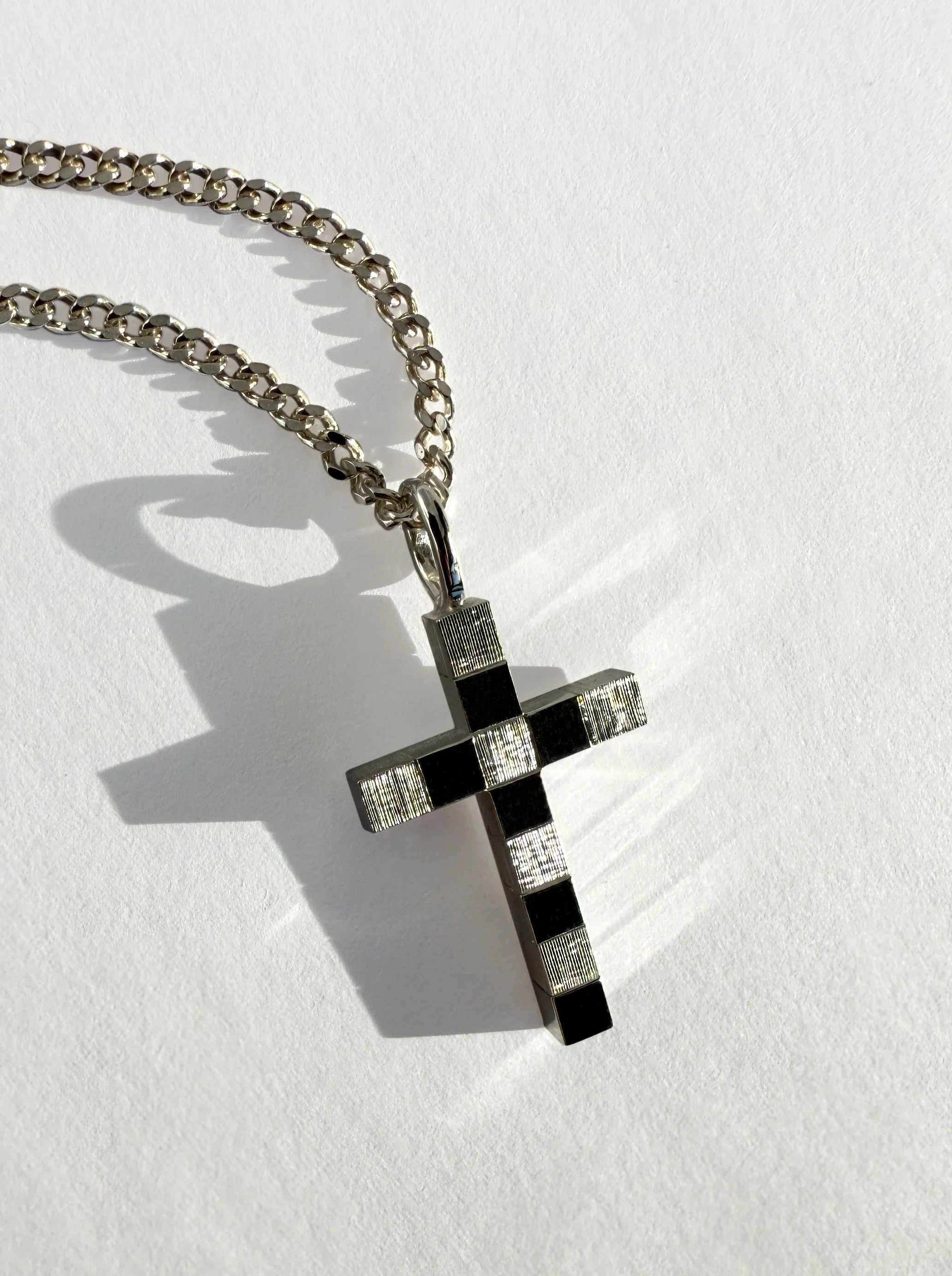 Silver chain necklace with a cross pendant that has black and silver striped pattern, lying on a white surface with a shadow cast by the sunlight.