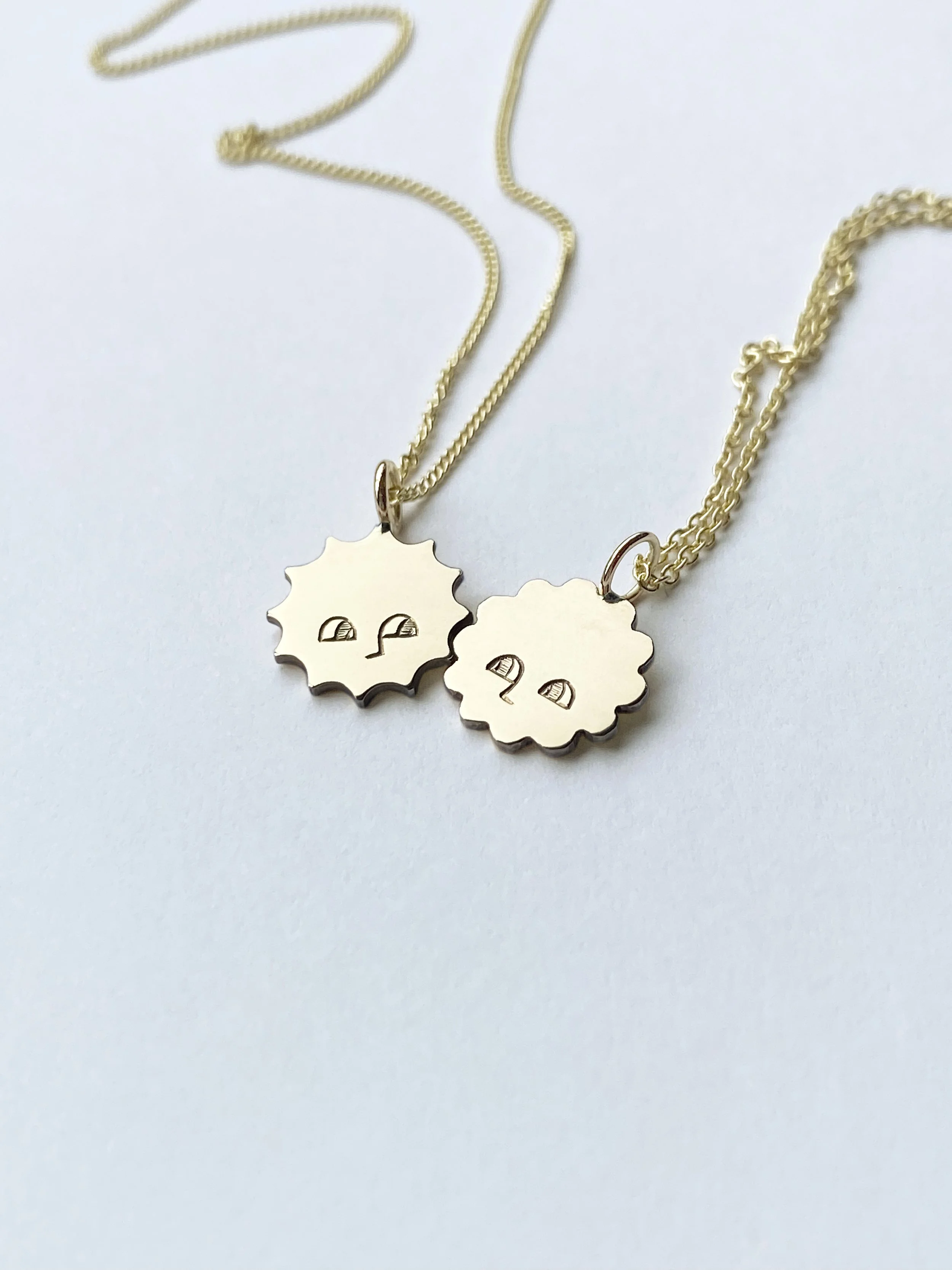Two gold necklaces with pendant charms resembling cartoon-style sun and moon faces on a white background.