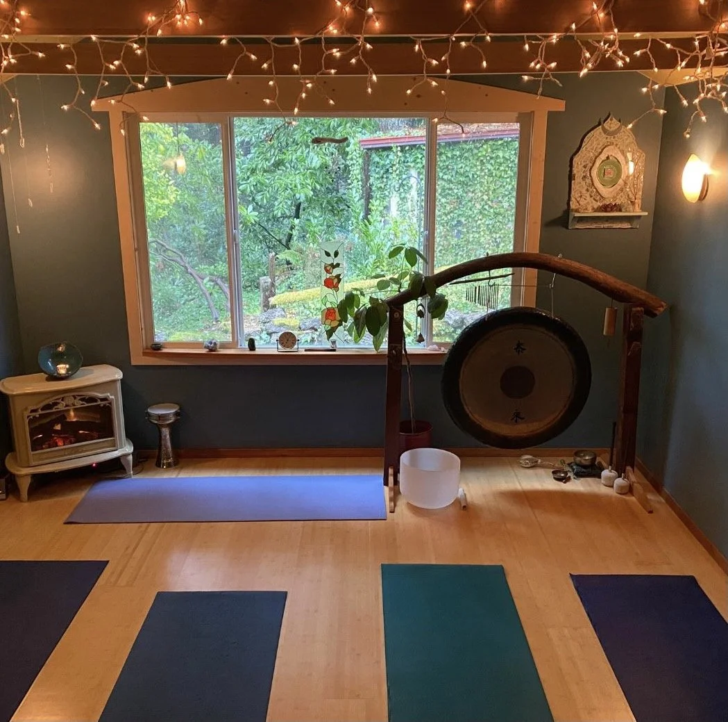 A cozy yoga studio with dim lighting and string lights. The room features several yoga mats on a wooden floor, a large window with a view of greenery outside, and a gong.