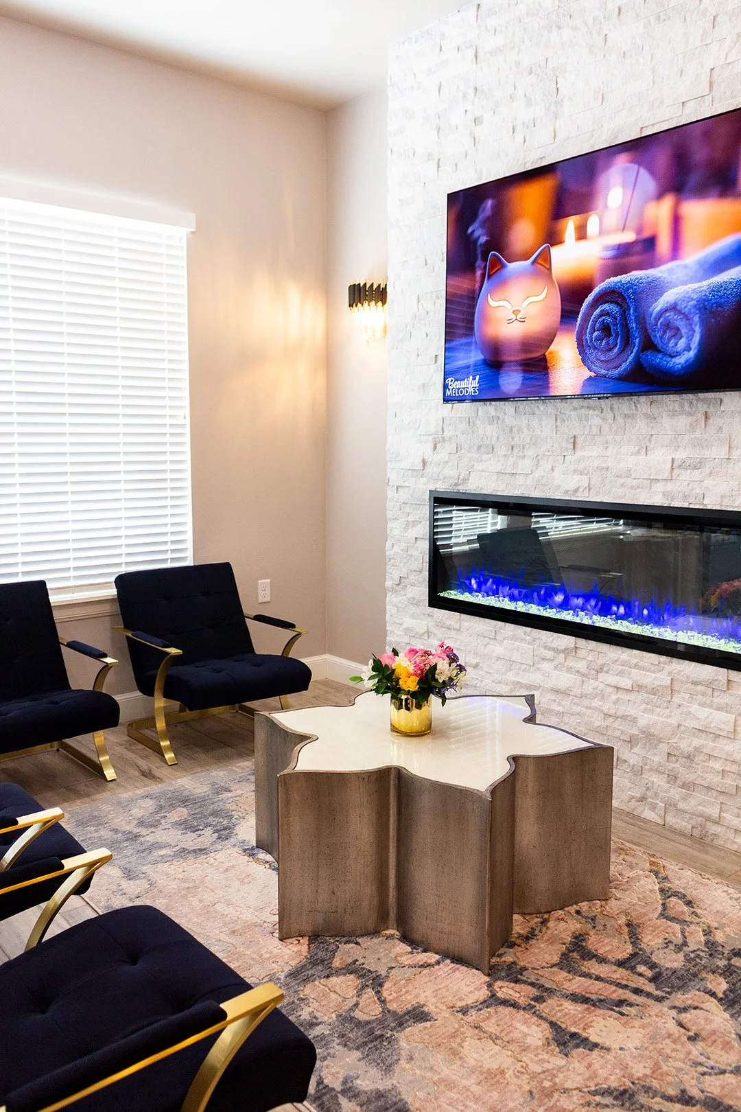 A modern living room with black armchairs with gold accents arranged around a white decorative coffee table with a flower arrangement. The room features a white brick wall with a mounted TV showing a relaxing scene and a built-in fireplace emitting blue flames. There is a window with blinds letting in natural light and a colorful rug.