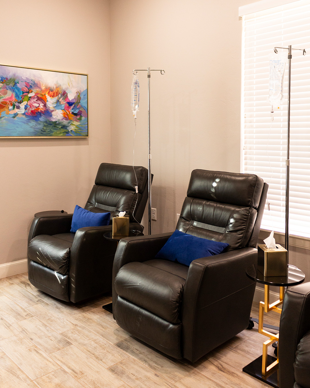 Two black leather recliner chairs with blue pillows, each with a side table holding tissue boxes and a gold decorative item, in a room with beige walls, a wooden floor, and a large window with blinds. IV stands with IV bags are positioned between the chairs.