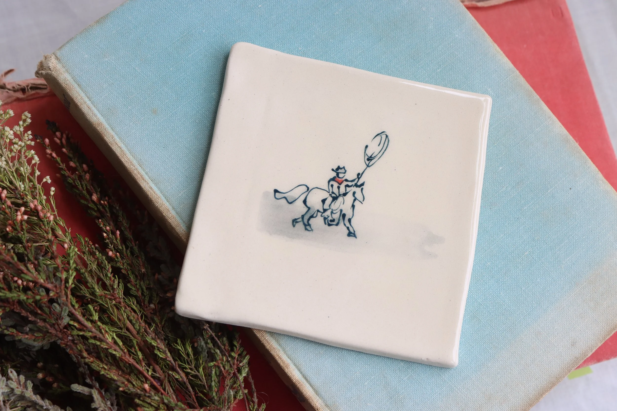 A white tile with a blue fine line illustration of a cowboy on a horse, throwing a lasso.