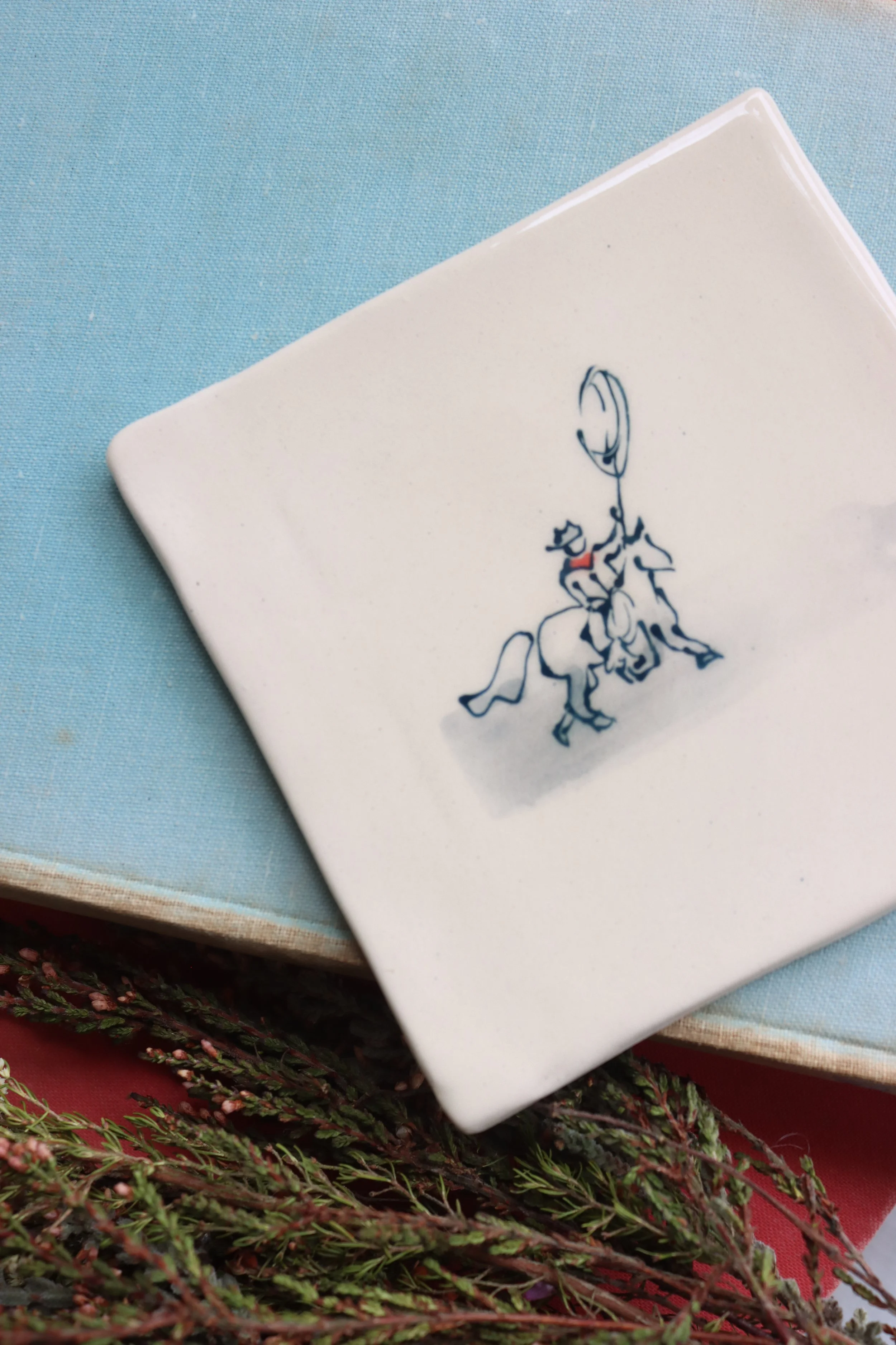 A white tile with a blue fine line illustration of a cowboy on a horse, throwing a lasso.