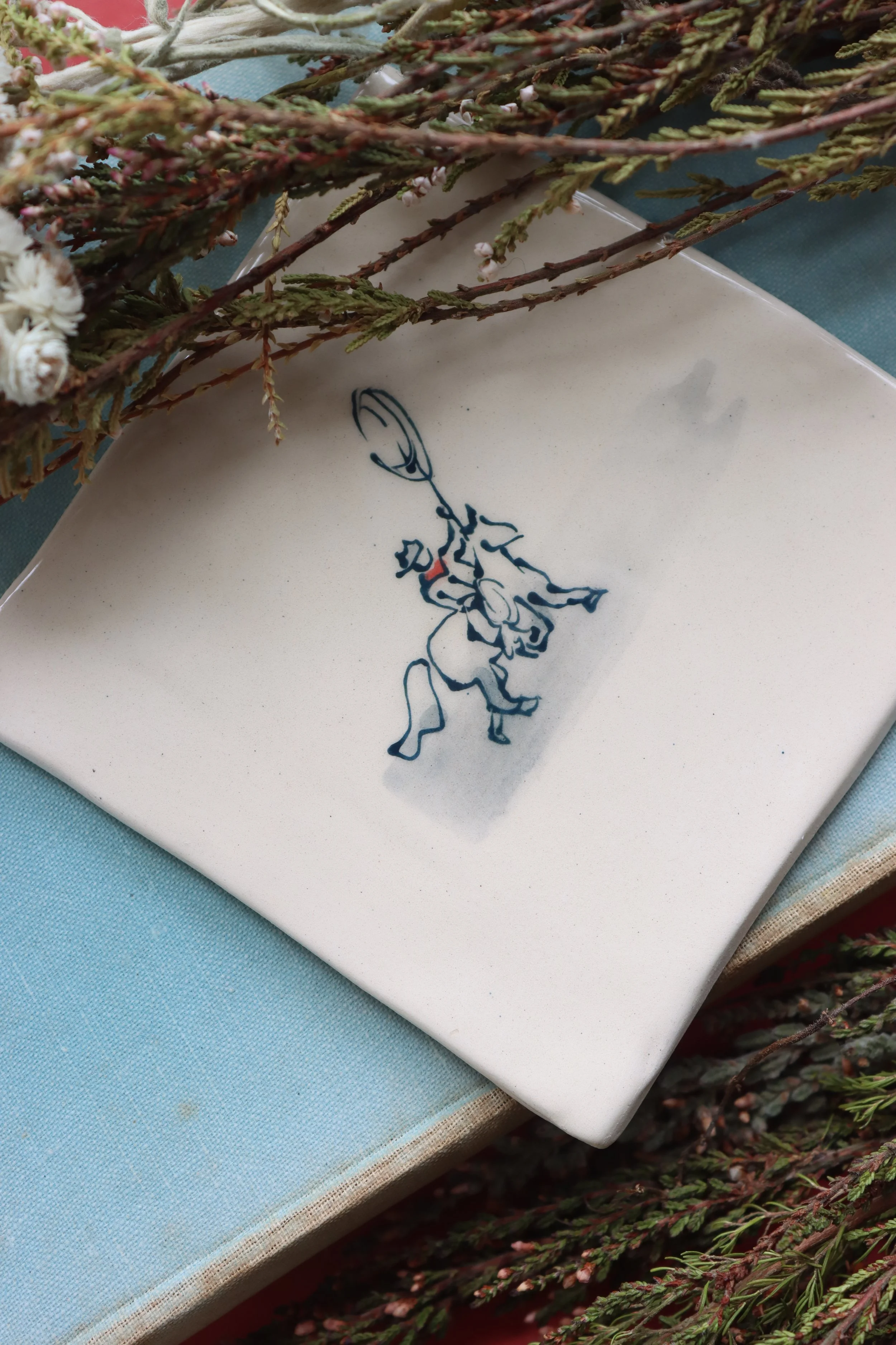 A white tile with a blue fine line illustration of a cowboy on a horse, throwing a lasso.