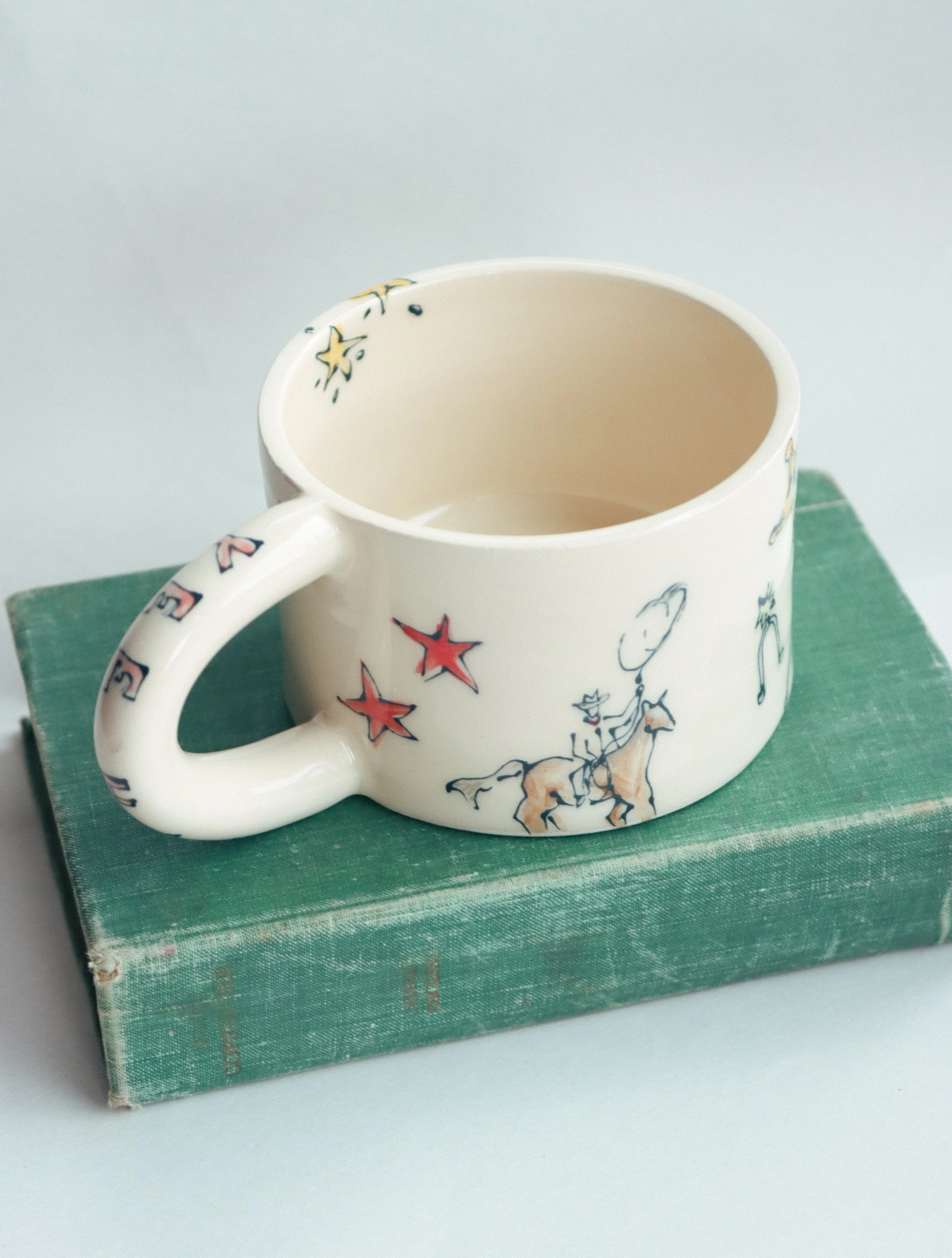 A cowboy themed illustrated mug, featuring a cowboy with a lasso on the side. It sits on a green book against a white background.