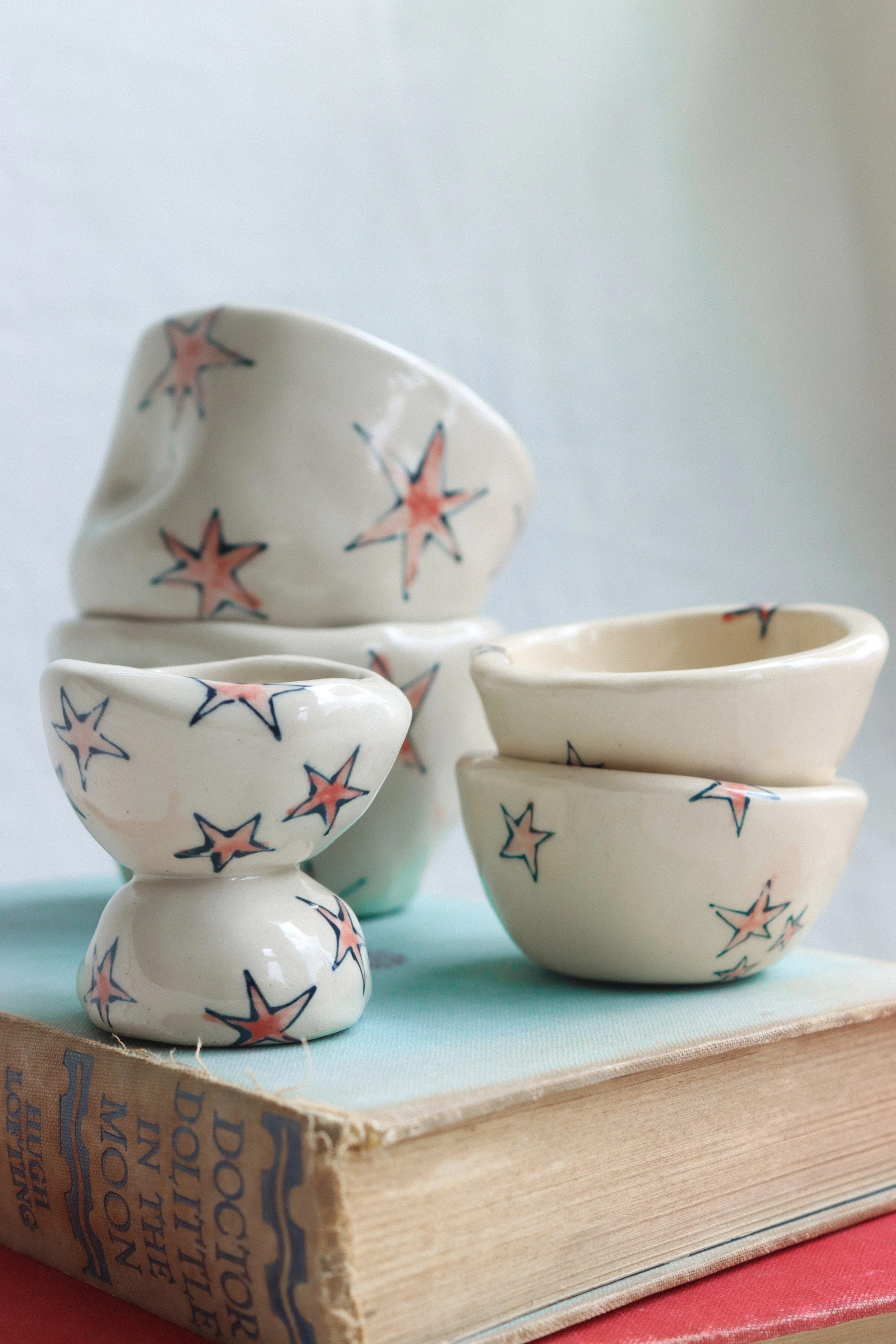 Two white ceramic cups stacked, two small dishes, and an eggcup arranged on a book. Each one is covered with red stars with blue outlines.