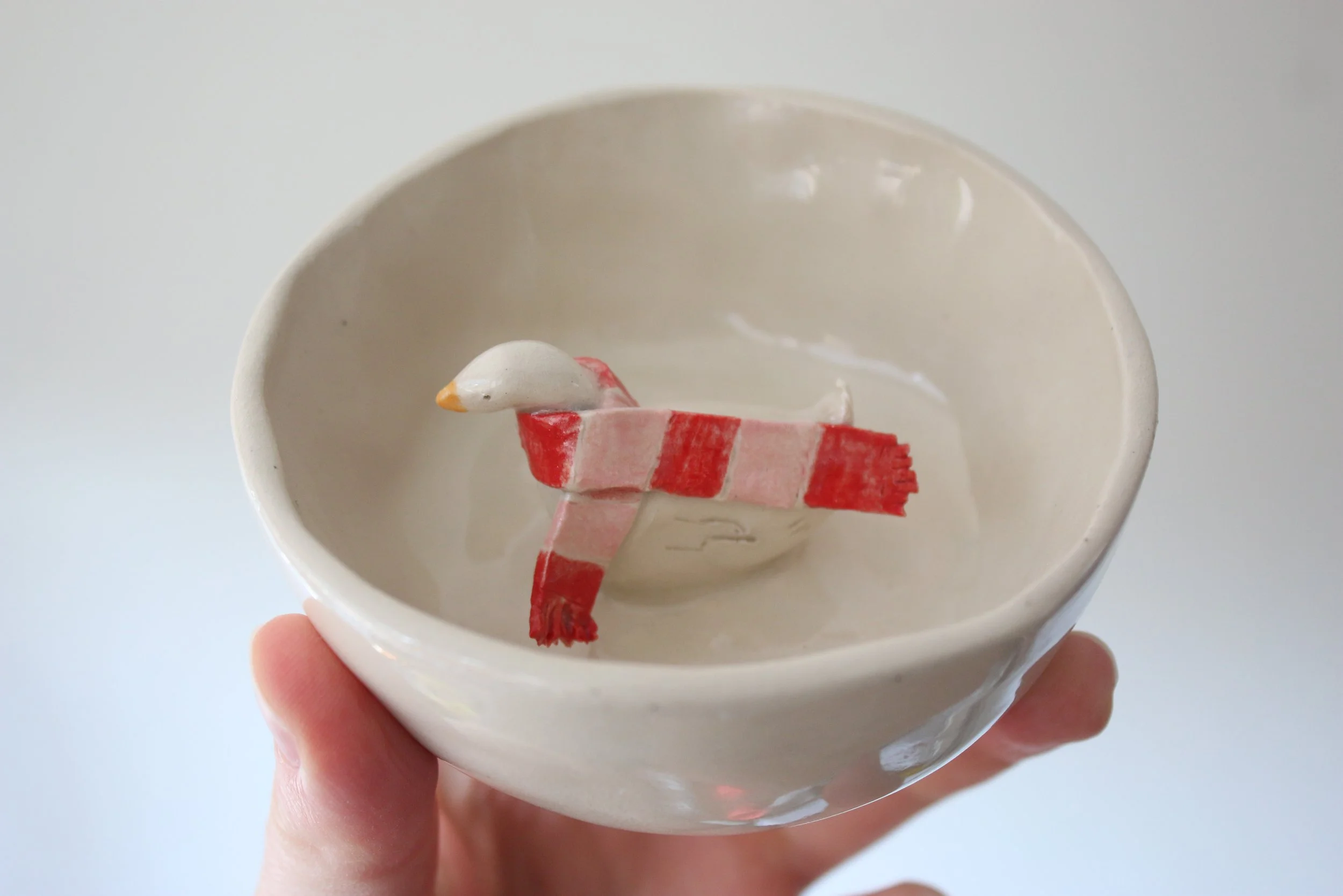 A small, off white, ceramic dish with a little duck in the middle. It is wearing a scarf.