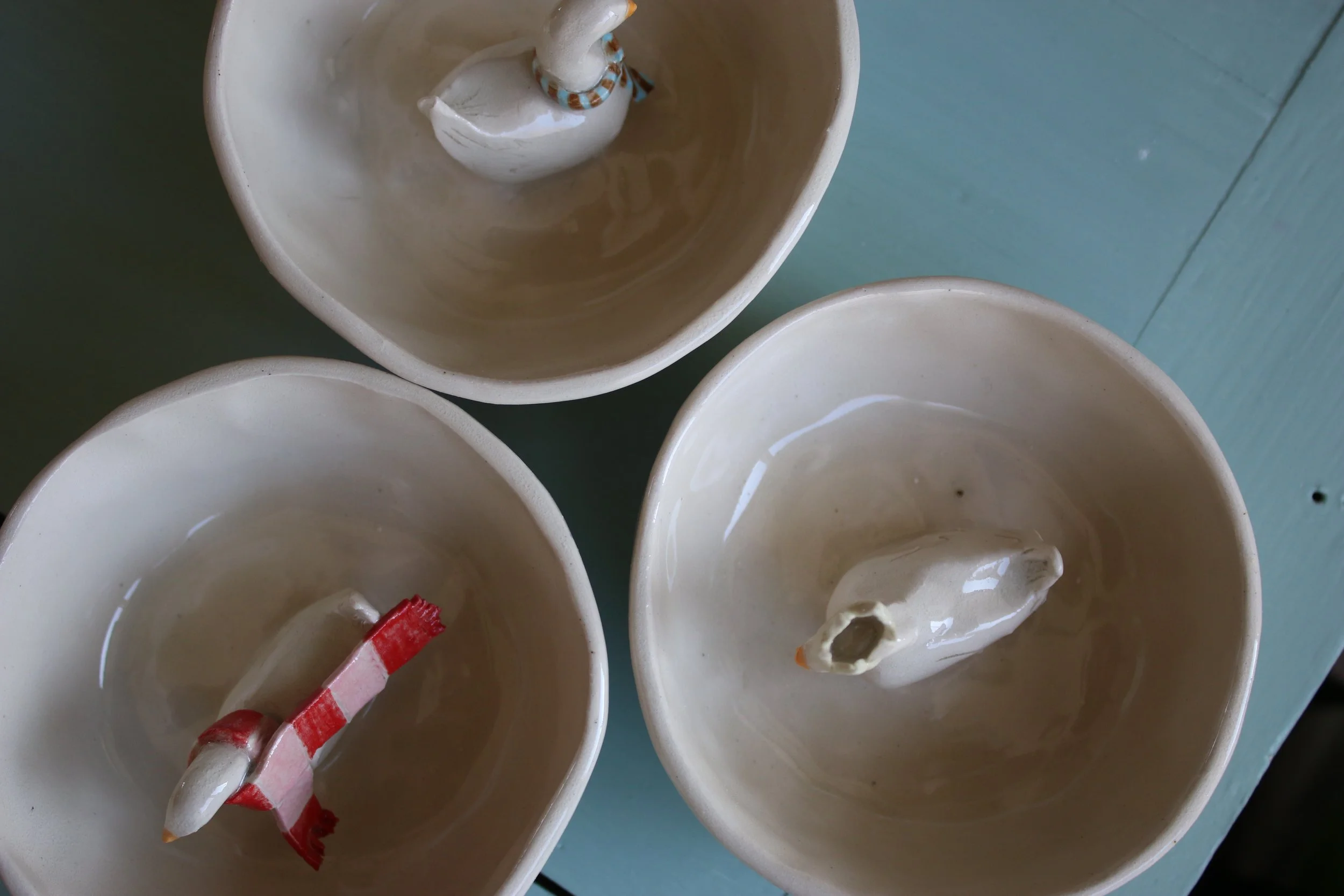 Three small, off white, ceramic dish with a little duck in the middle. They are each respectively wearing a crown, tie, and a scarf.