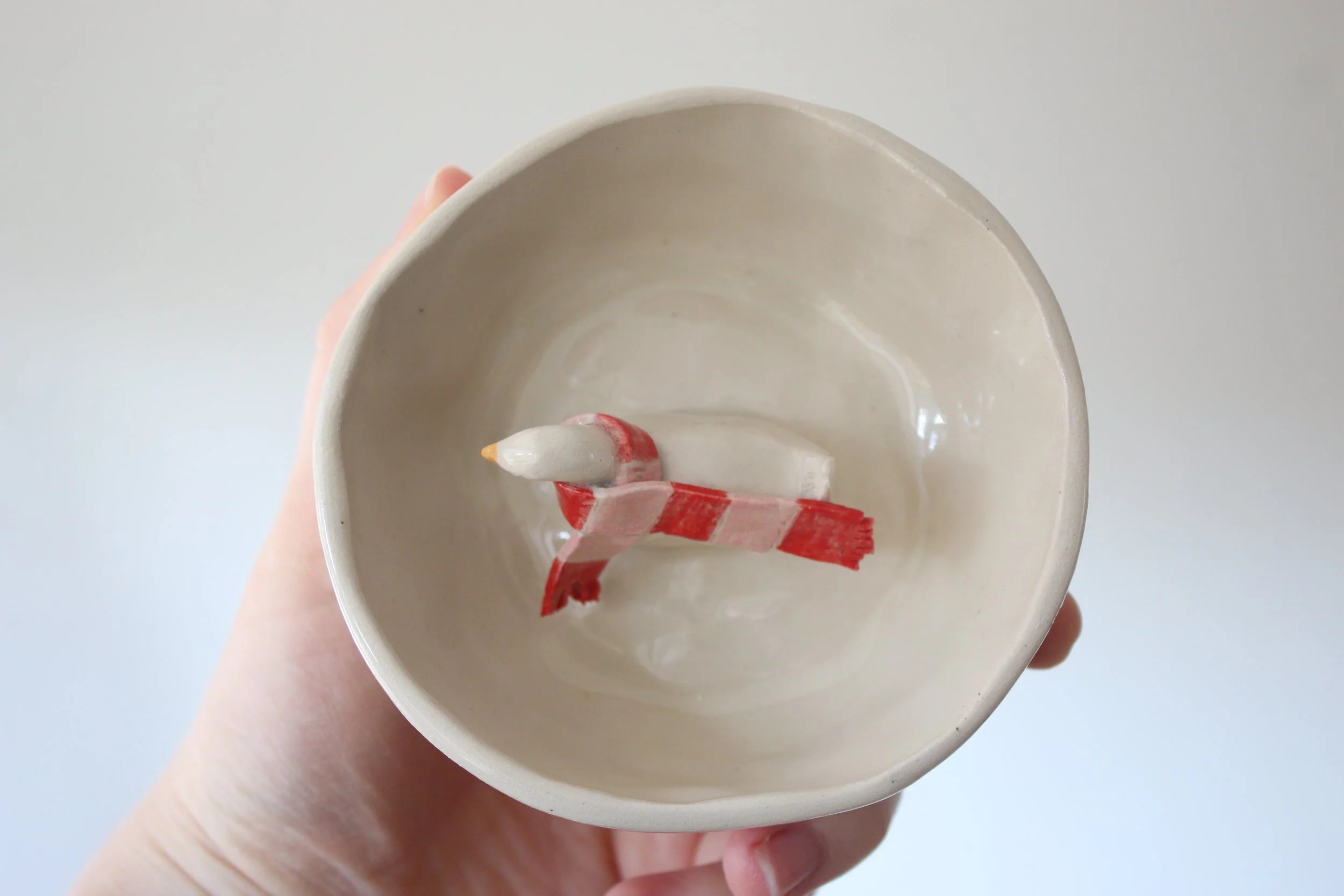 A small, off white, ceramic dish with a little duck in the middle. It is wearing a scarf.