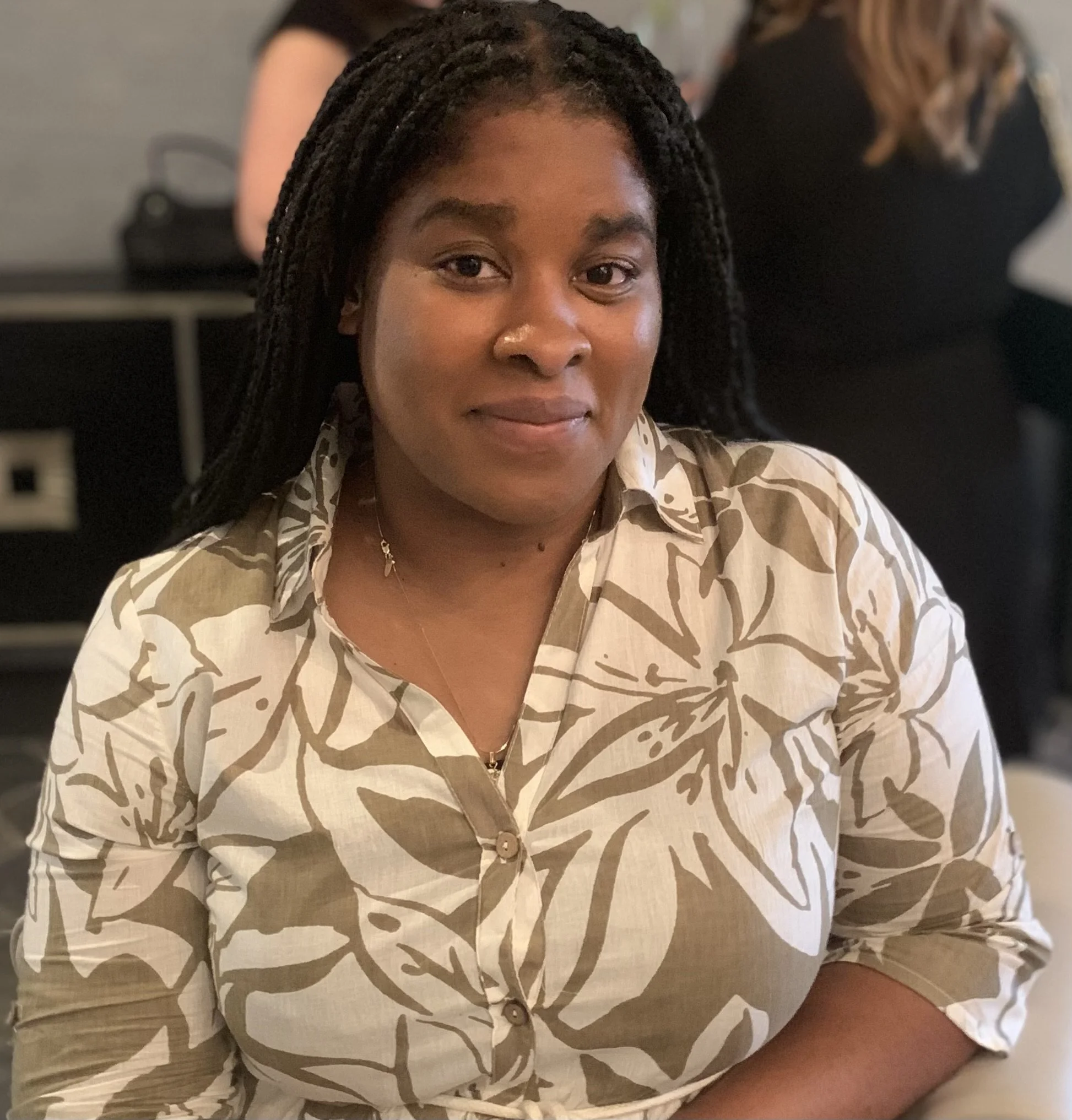 A woman (Shereka Binnie) with dark braided hair wearing a beige and white floral patterned shirt sitting at a table indoors.