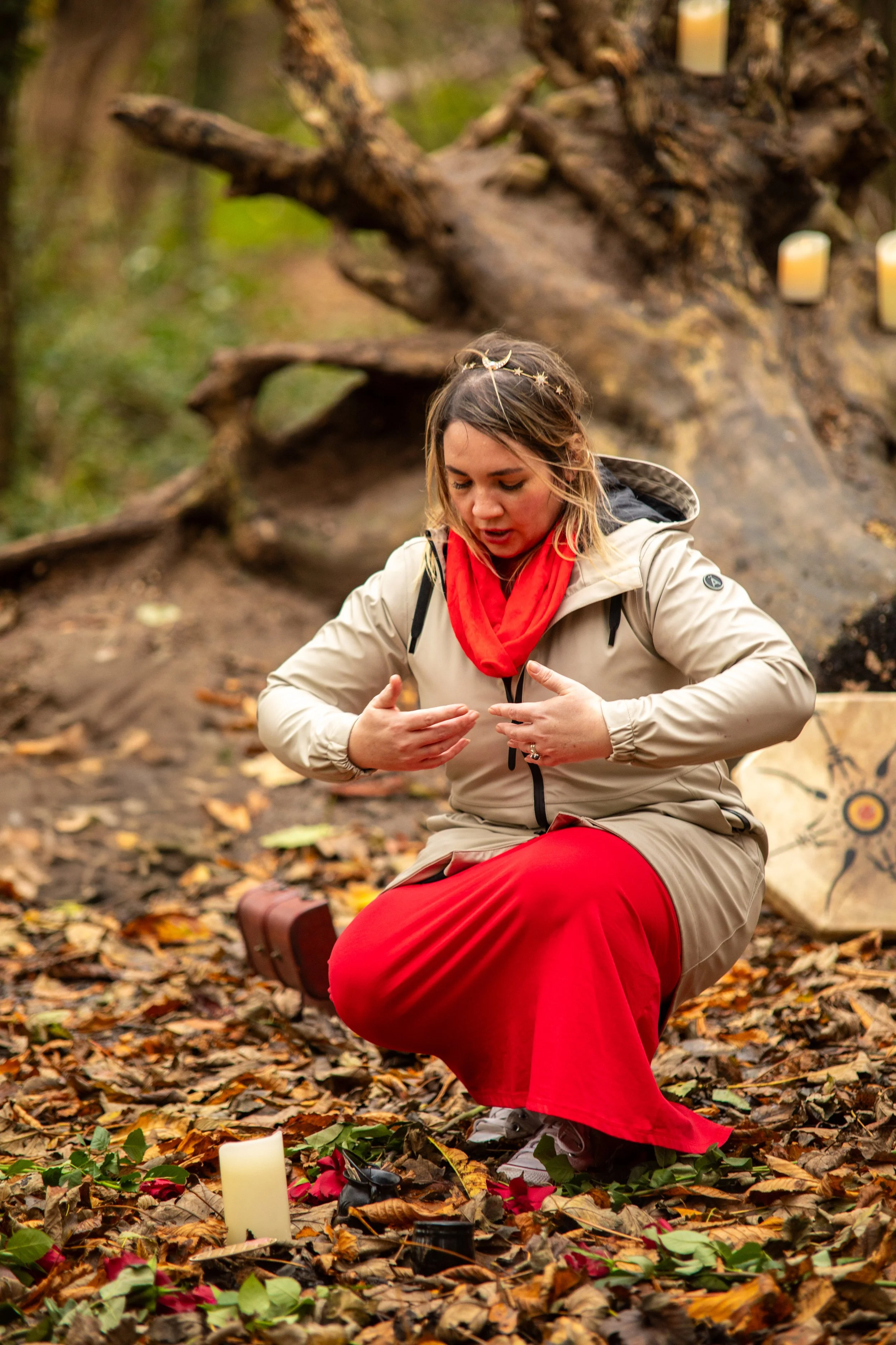 A woman dressed in a beige jacket and red skirt is kneeling on fallen leaves in a forest, with a large tree base and candles in the background, and appears to be participating in a ritual or ceremony.