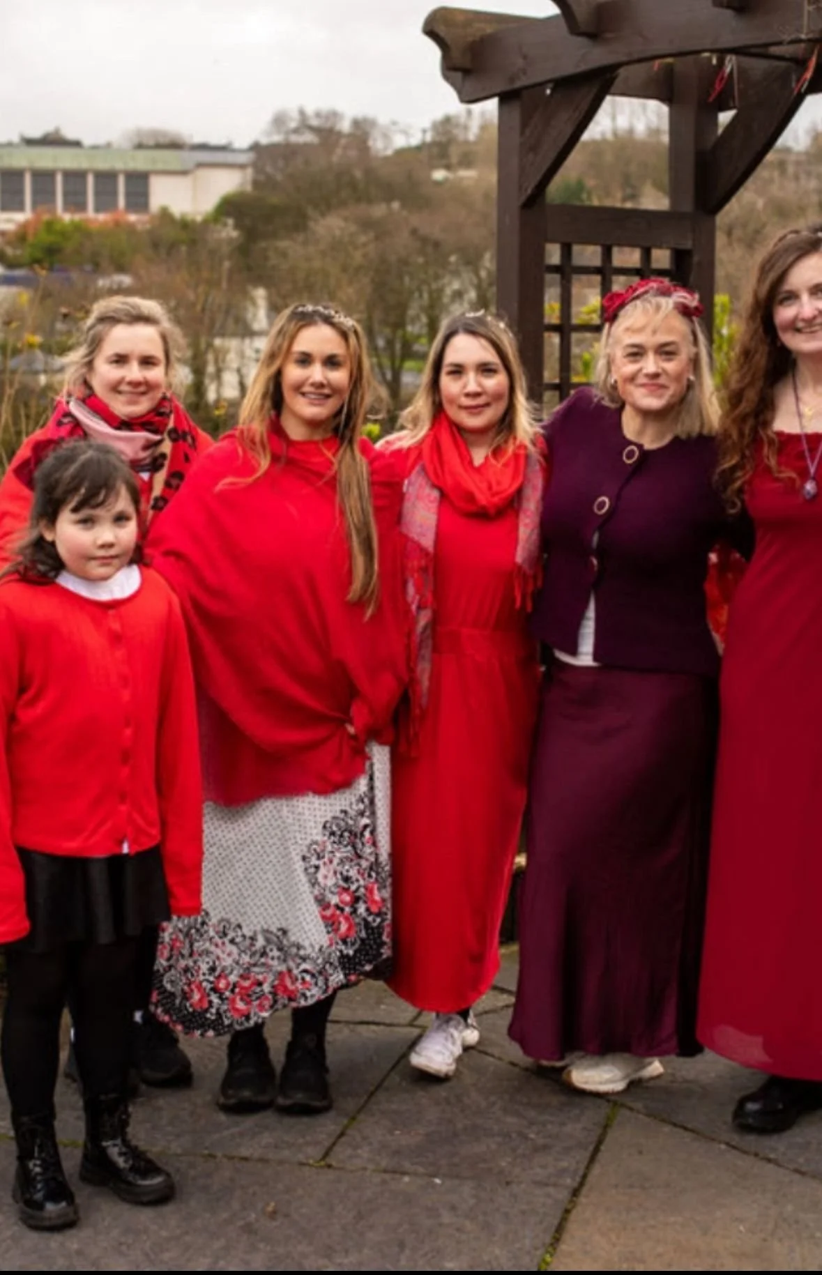 Group of six women and one girl standing outdoors, dressed in red and dark clothing. They are smiling, with trees and buildings in the background.