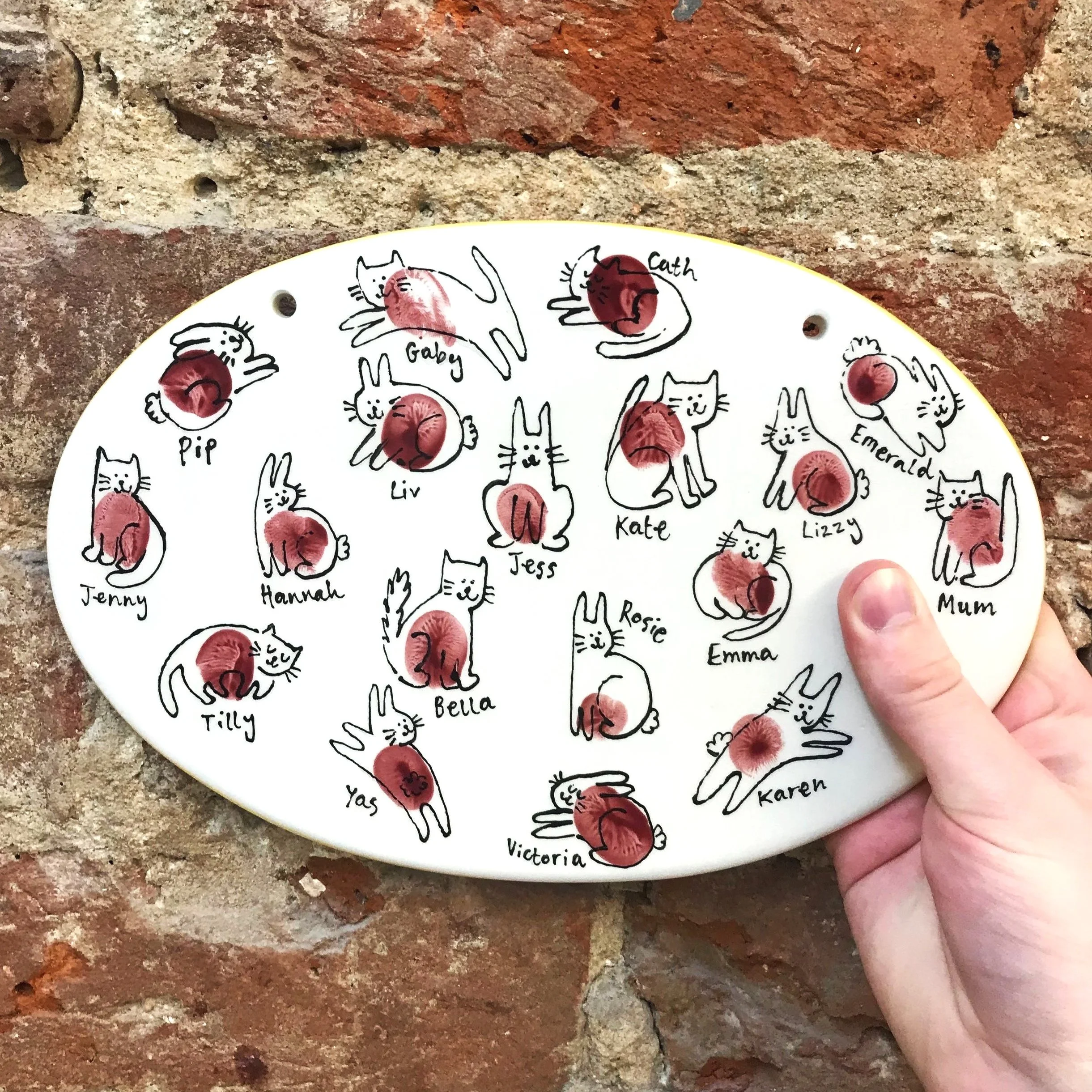 A decorative ceramic plate with illustrations of cats in various poses, each labeled with a name. Some cats have lipstick marks or eyeshadow, and a person's finger is holding the plate, which is against a brick wall.