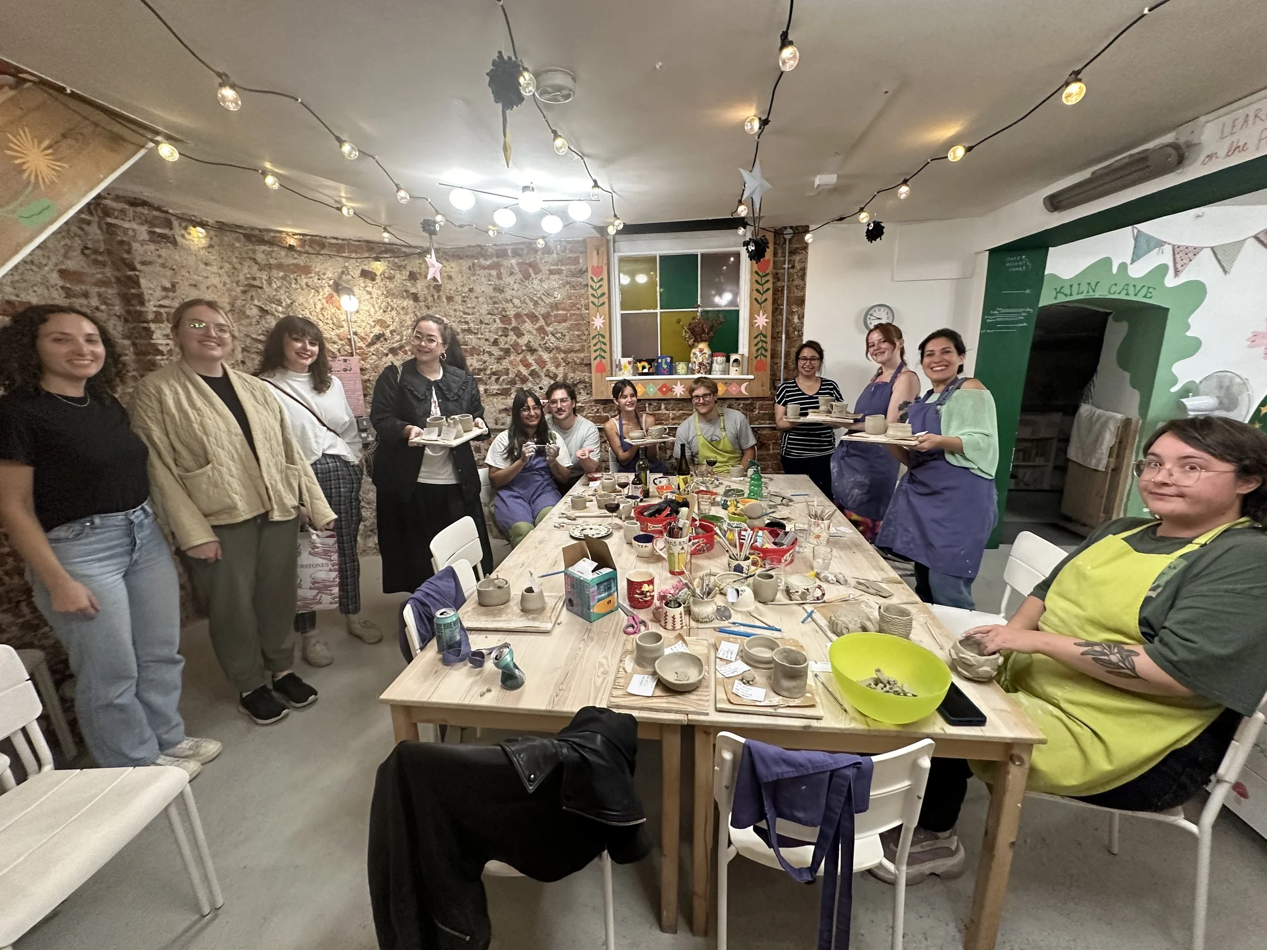 A group of people gathered around a table for a pottery painting activity in a cozy, decorated room with string lights and brick walls.