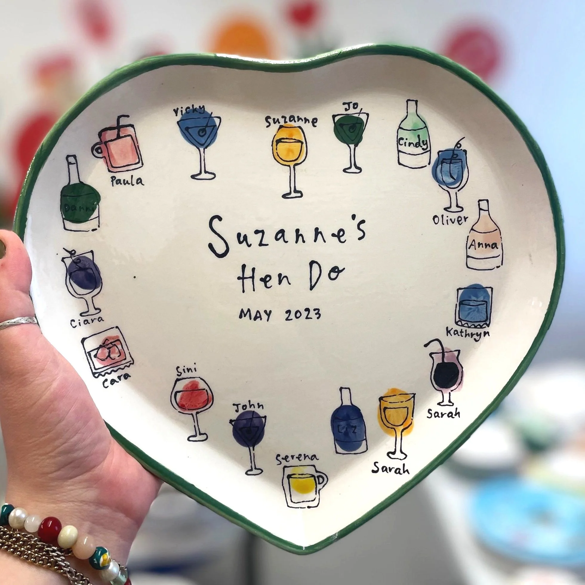 Decorative heart-shaped plate with colorful illustrations of drinks and names around the edge, and text in the center that reads "Suzanne's Hen Do MAY 2023."