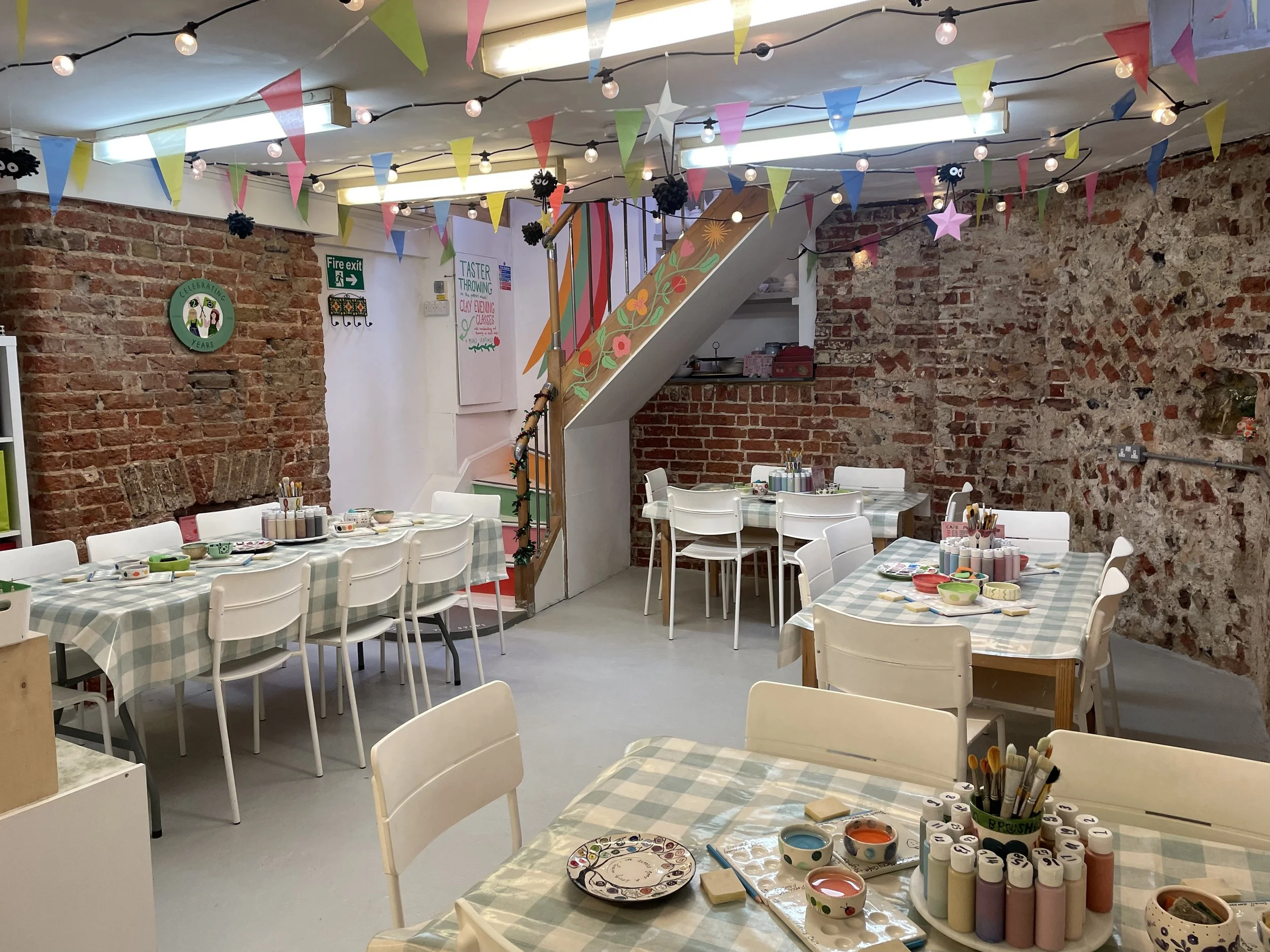A cozy craft room decorated with colorful bunting, star and heart shaped banners, string lights, and paper flowers. There are several tables covered with checkered tablecloths, set up with paints, brushes, and craft supplies. Exposed brick walls add a rustic touch to the space.