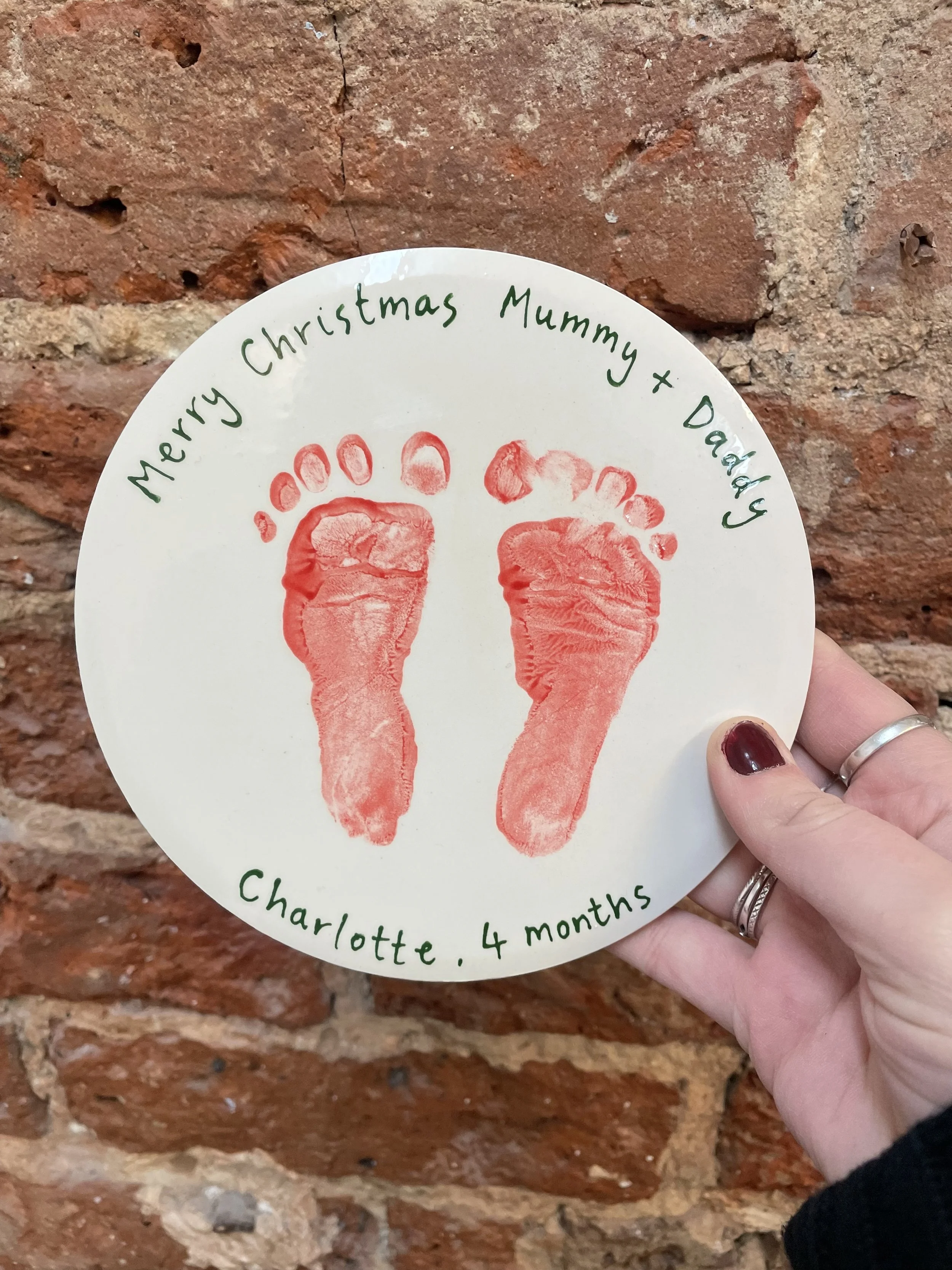 A circular keepsake with red handprints of a baby or young child, along with handwritten green text wishing a Merry Christmas to Mummy and Daddy, from Charlotte at four months old, against a brick wall background.