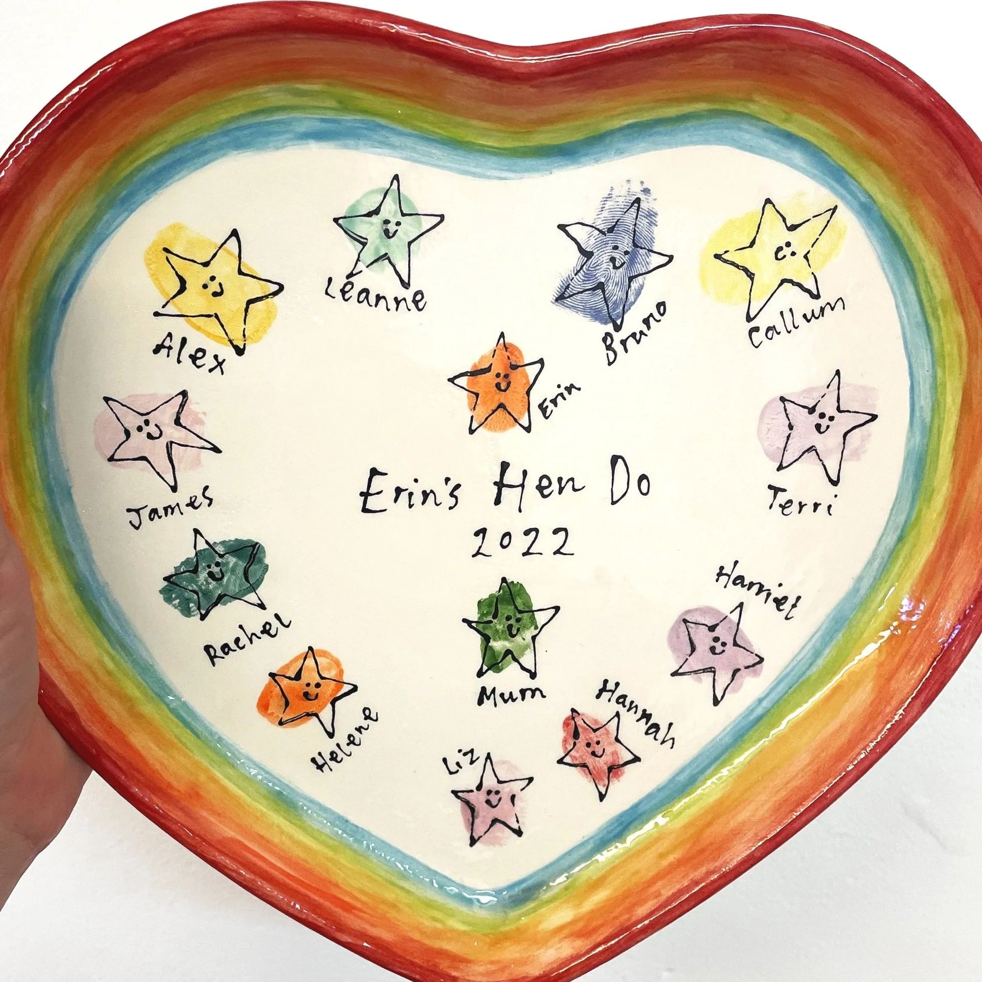 Colorful heart-shaped ceramic plate with hand-drawn smiling stars in various colors and names around the edge, and 'Erin's Hen Do 2022' written in the center.
