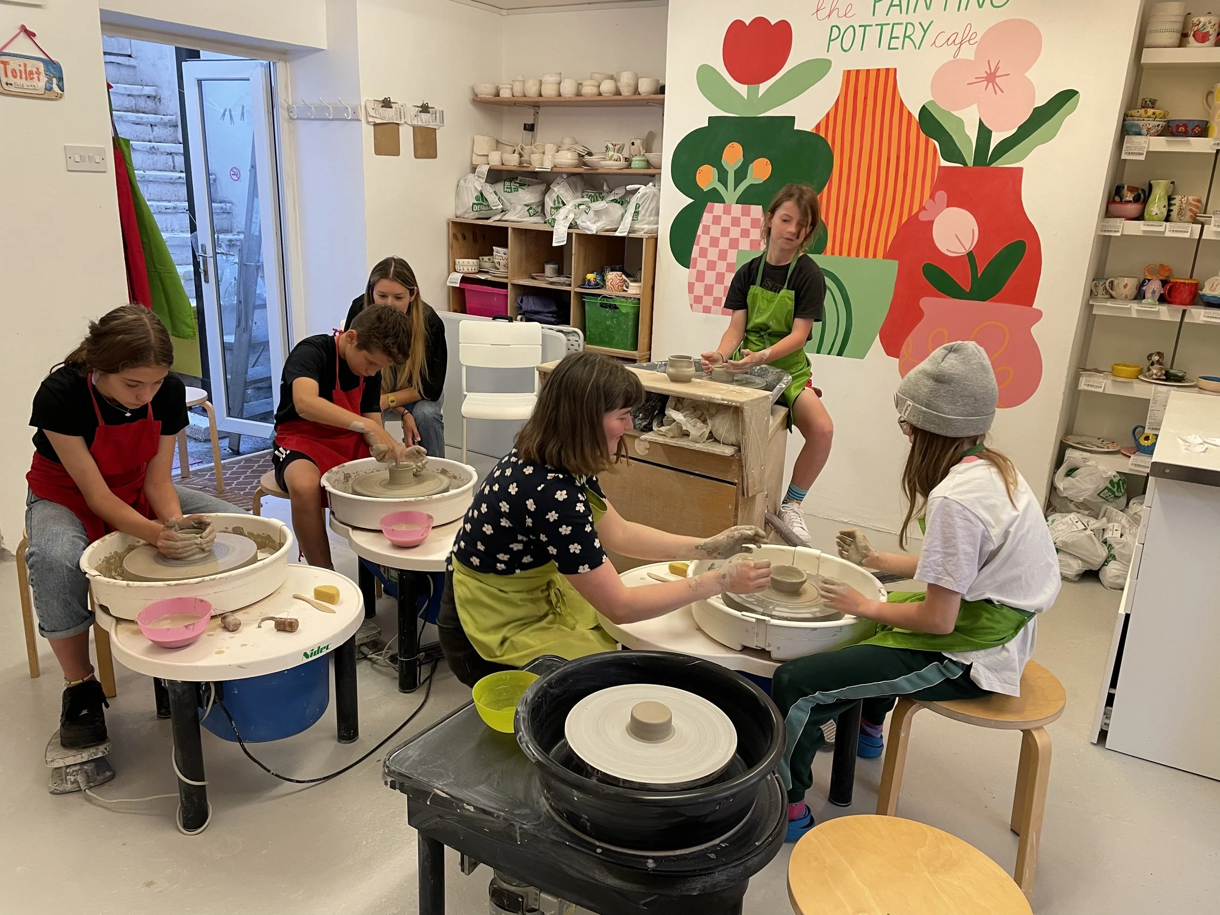 Children participating in a pottery class, working on pottery wheels in an art studio with colorful wall artwork and shelves of ceramics.