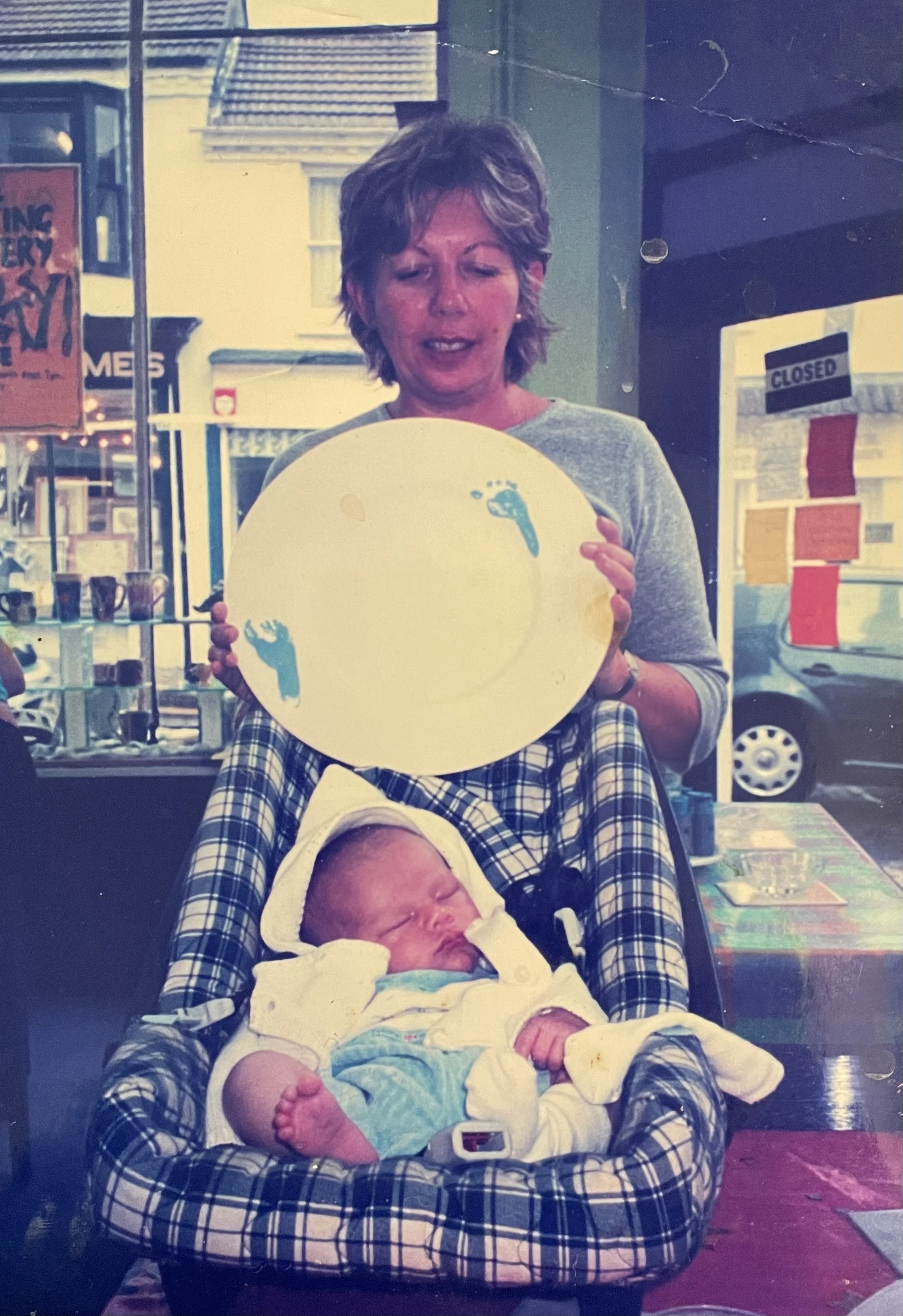 A woman with short hair holding a decorative plate with blue footprints, standing behind a baby in a car seat with a hood, who appears to be sleeping.
