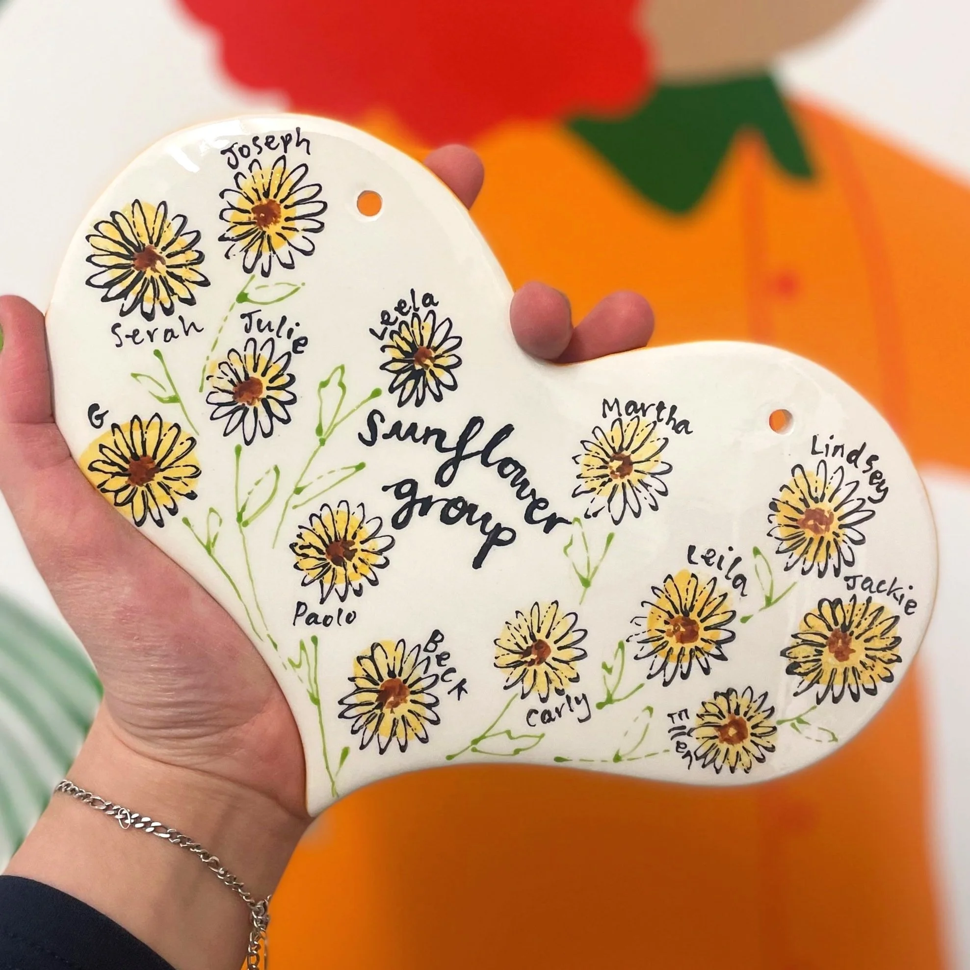 Hand holding a ceramic plaque with daisies and names of group members, reading 'Sunflower Group'. There is a pumpkin decoration in the background.