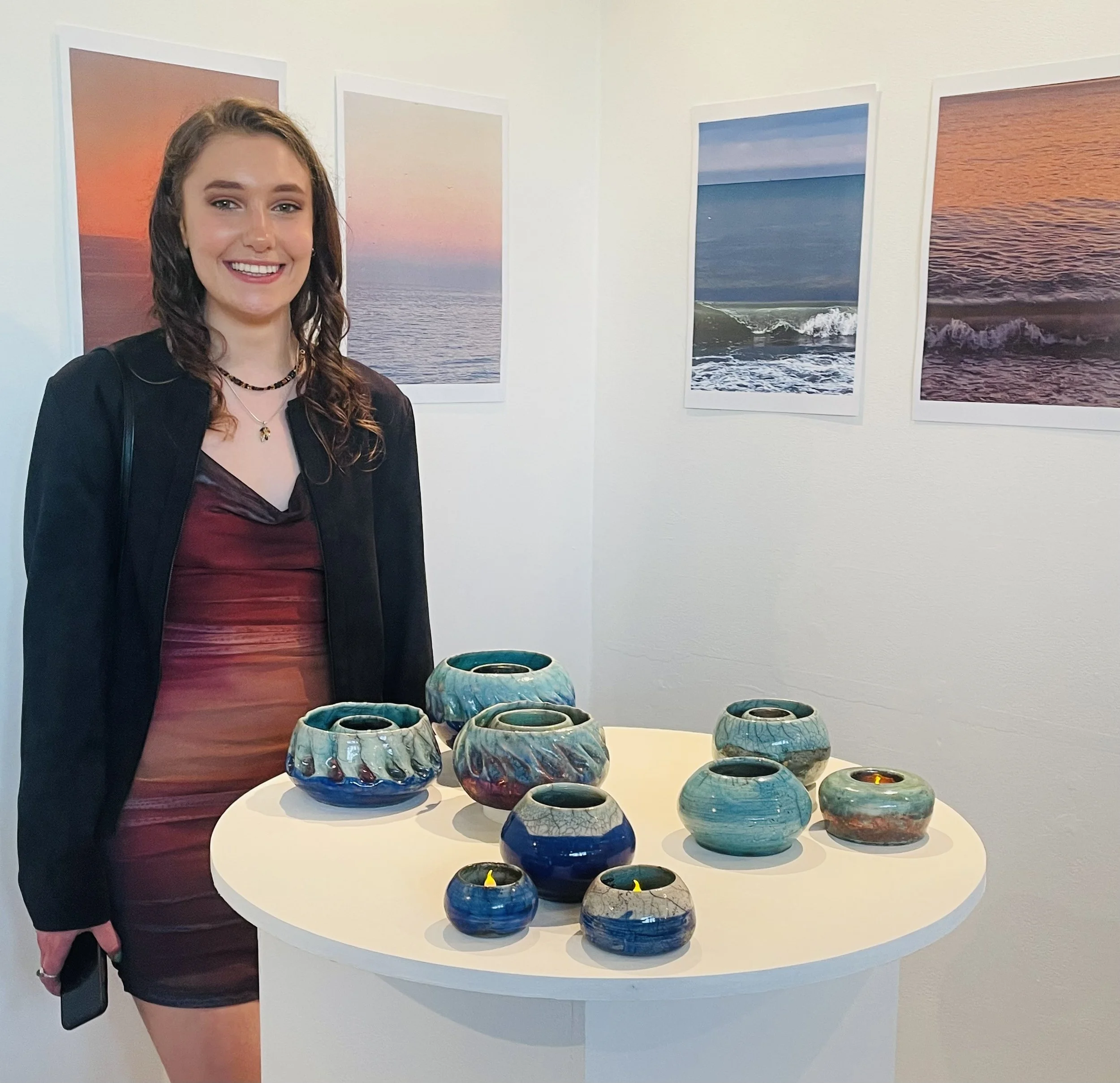 A young woman with wavy brown hair wearing a black jacket and a maroon and pink dress, standing next to a display table with colorful ceramic pots, with seascape photographs on the white gallery walls behind her.