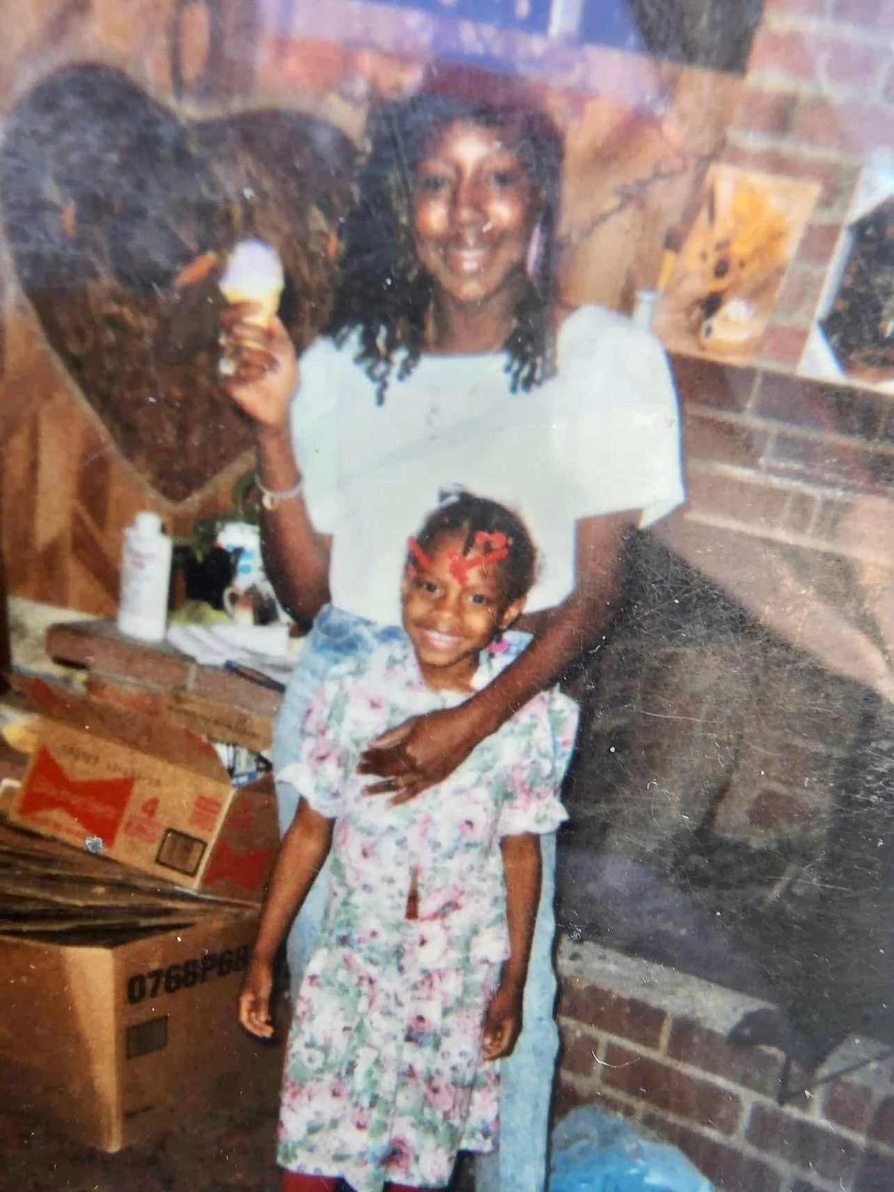 A woman with dark curly hair smiling and holding a spray bottle, standing behind a young girl with braided hair and a colorful dress, in a room with brick walls and various items and boxes around.
