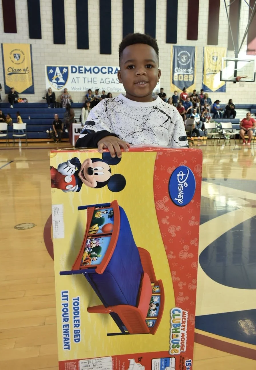 Vegas Black Pride® 2018 image: A young child standing in a gymnasium holding a Mickey Mouse Clubhouse toddler bed box, with a basketball hoop and banners in the background.