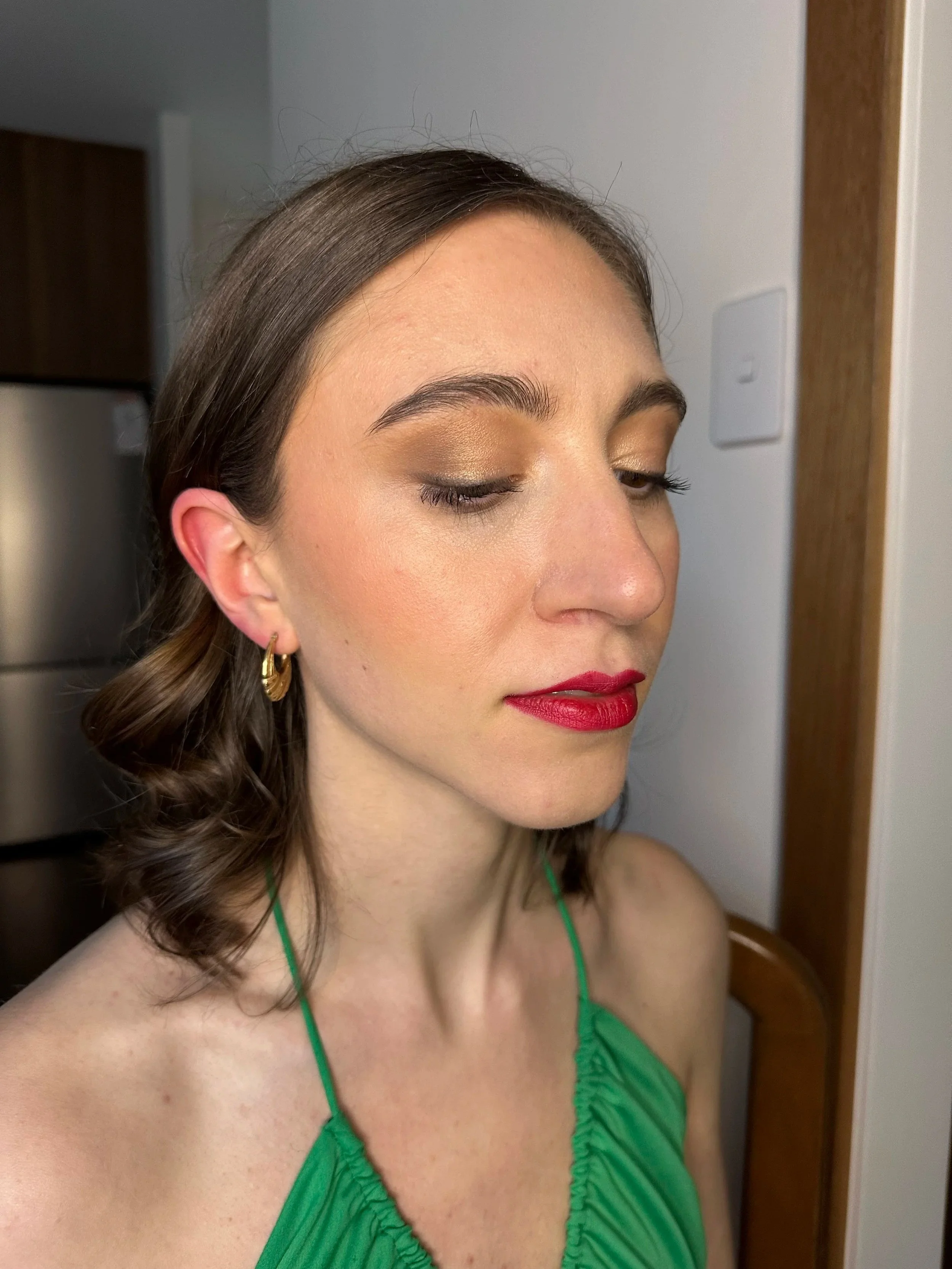 Close-up of a woman with makeup, wearing a green dress with thin straps, gold hoop earrings, and red lipstick, standing indoors with a neutral background.