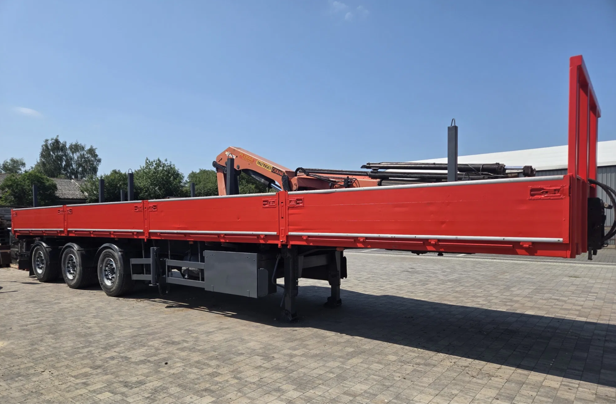 Red flatbed truck with a crane attachment parked on a cobblestone surface, with trees and a building in the background under a clear blue sky.
